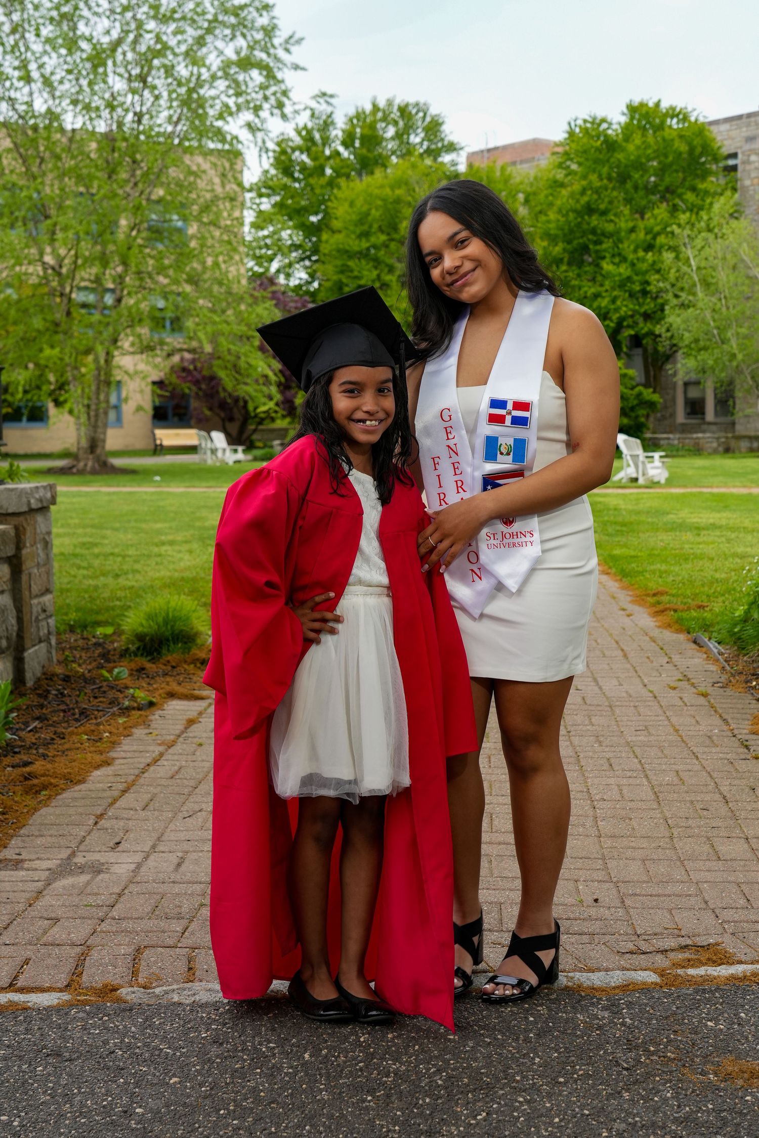 Two young women pose outdoors in graduation attire, one in red cap and gown, the other in a white dress with sash.