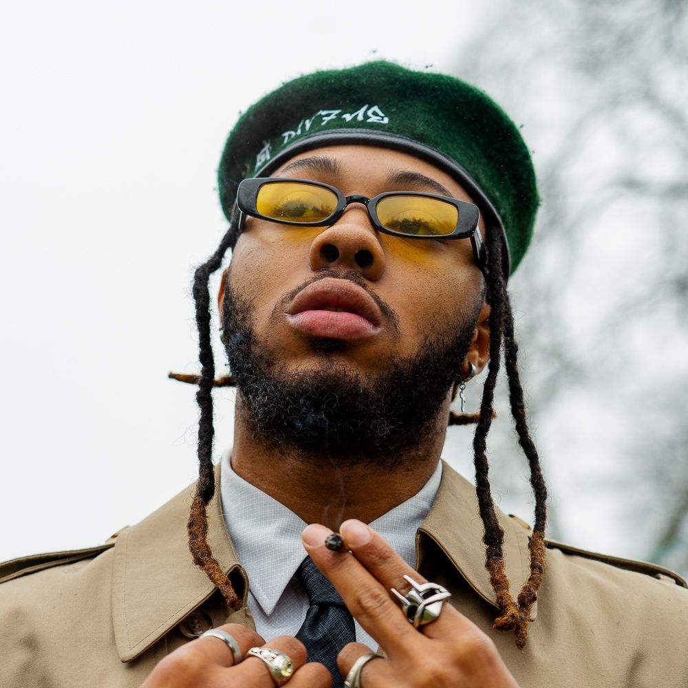Young man with dreadlocks wearing yellow tinted sunglasses, green beret, trench coat, and rings poses outdoors.