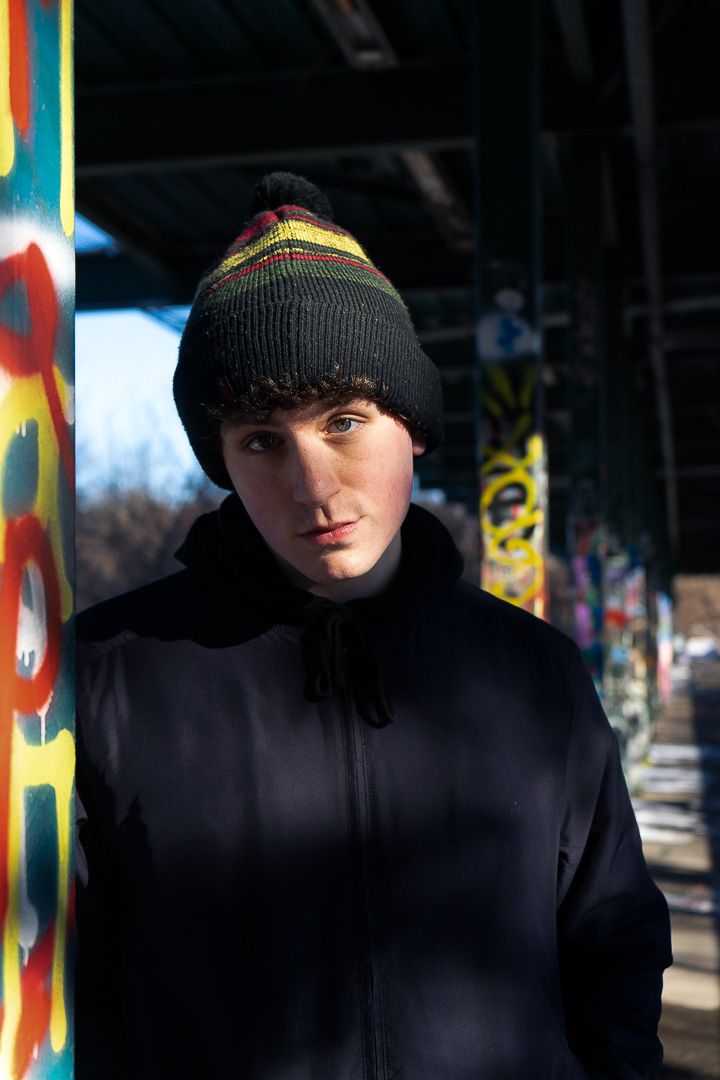 Young man in black coat and striped knit beanie poses near colorful graffiti-covered columns outdoors.