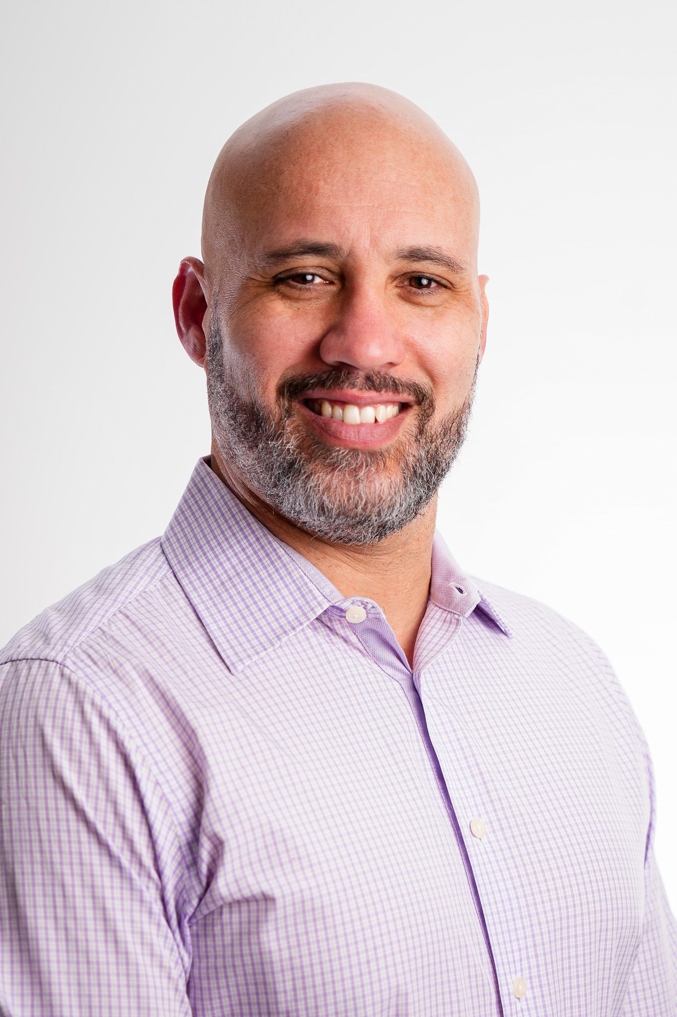 Smiling bald man with a gray beard wearing a light purple checkered shirt against a white background.