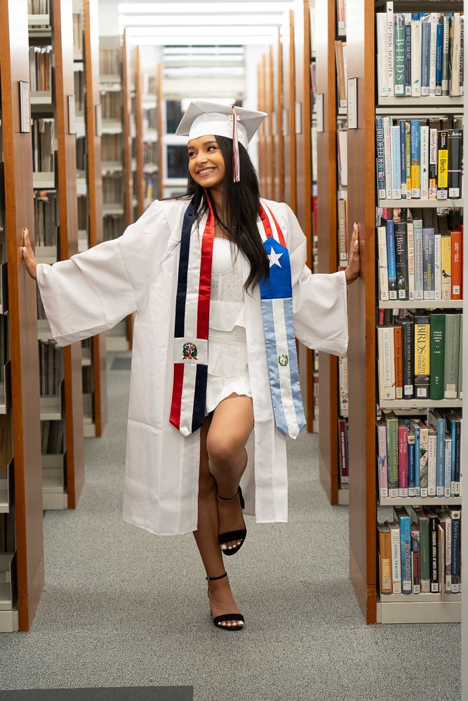 Smiling graduate in white cap and gown with flag sashes posing between library bookshelves.