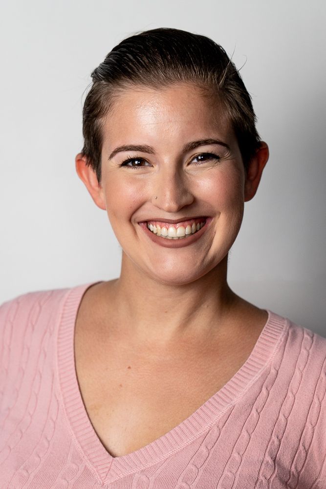 Smiling young woman with dark hair wearing a pink cable-knit V-neck sweater against a gray background.
