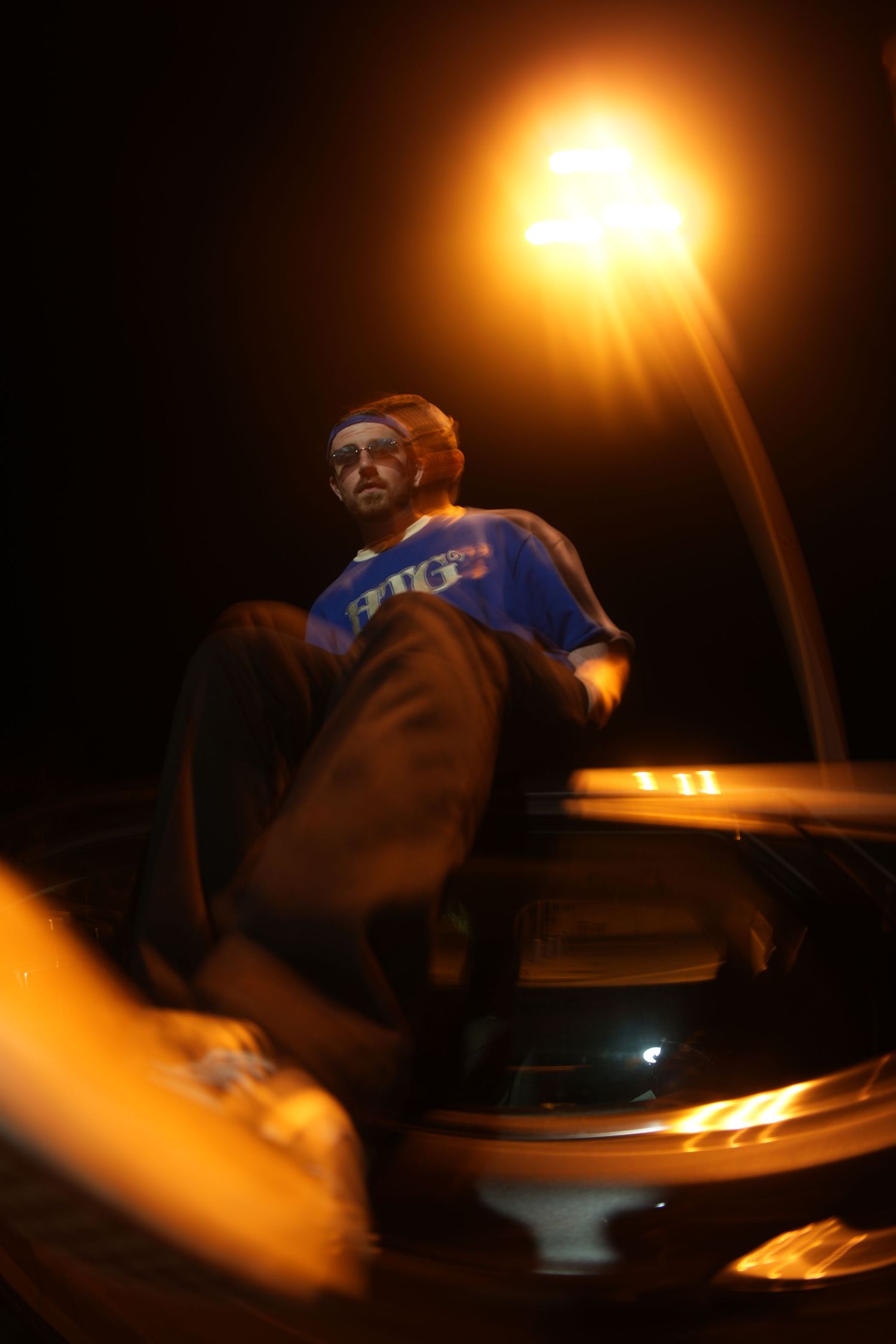 Man in blue jersey sitting on car hood under street lamp at night.
