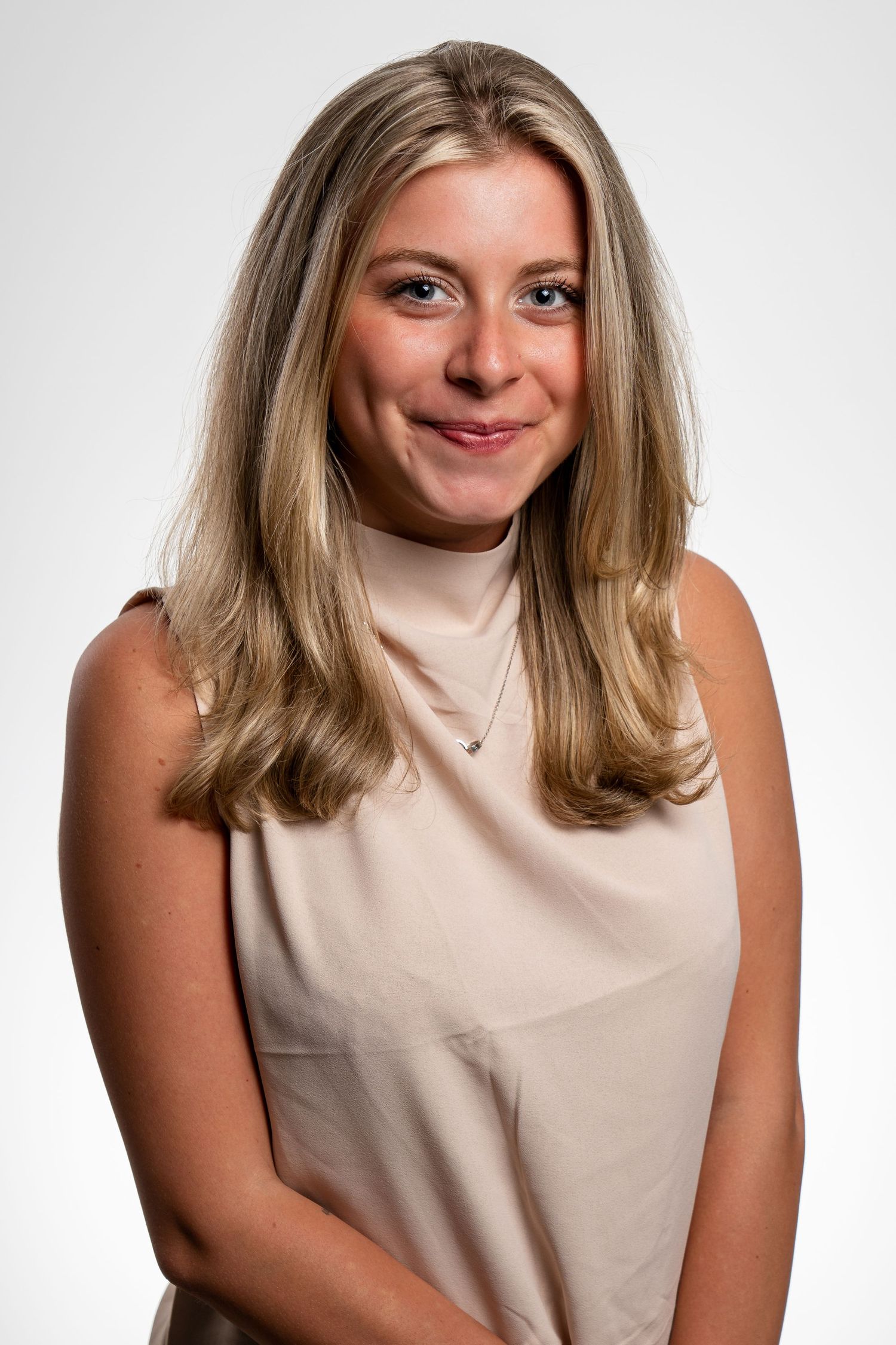 Young blonde woman smiling in a sleeveless beige top, professional headshot on white background.