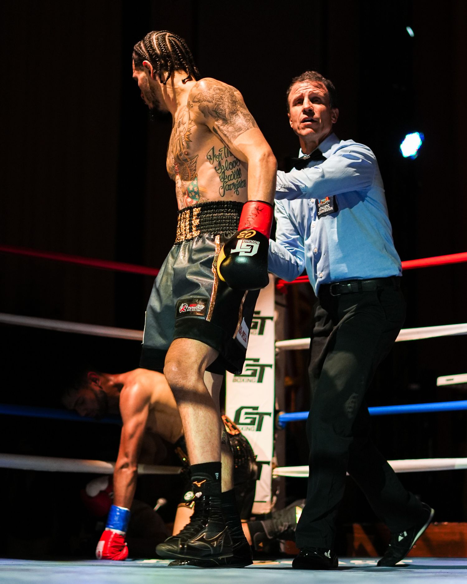 Referee guides tattooed boxer in professional boxing ring during match.