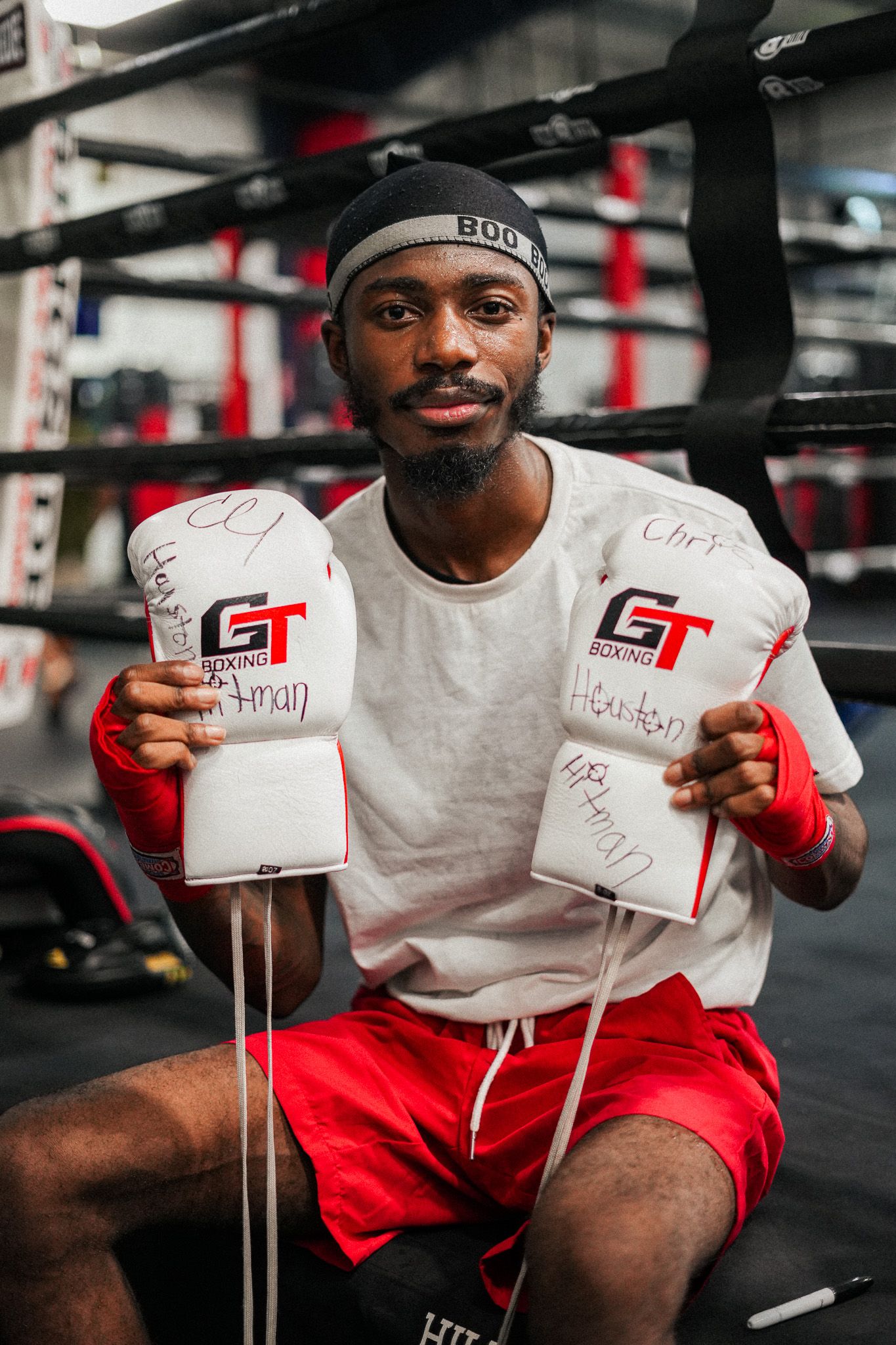 Boxer in red shorts holds up signed boxing gloves in a gym setting.