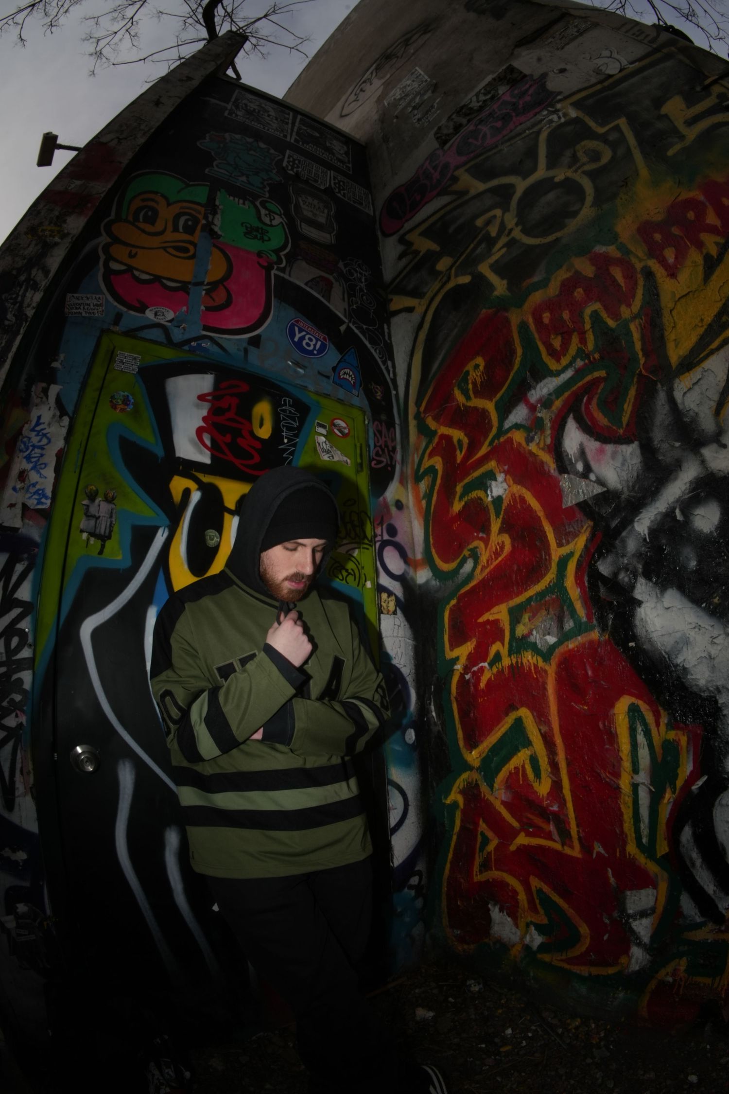 Man in green jacket and black hoodie standing against colorful graffiti wall in urban setting.