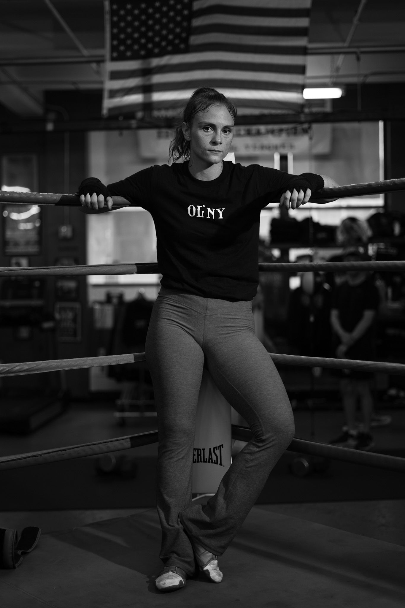 Athlete in OFNY sweatshirt poses against boxing ring ropes with American flag in background.