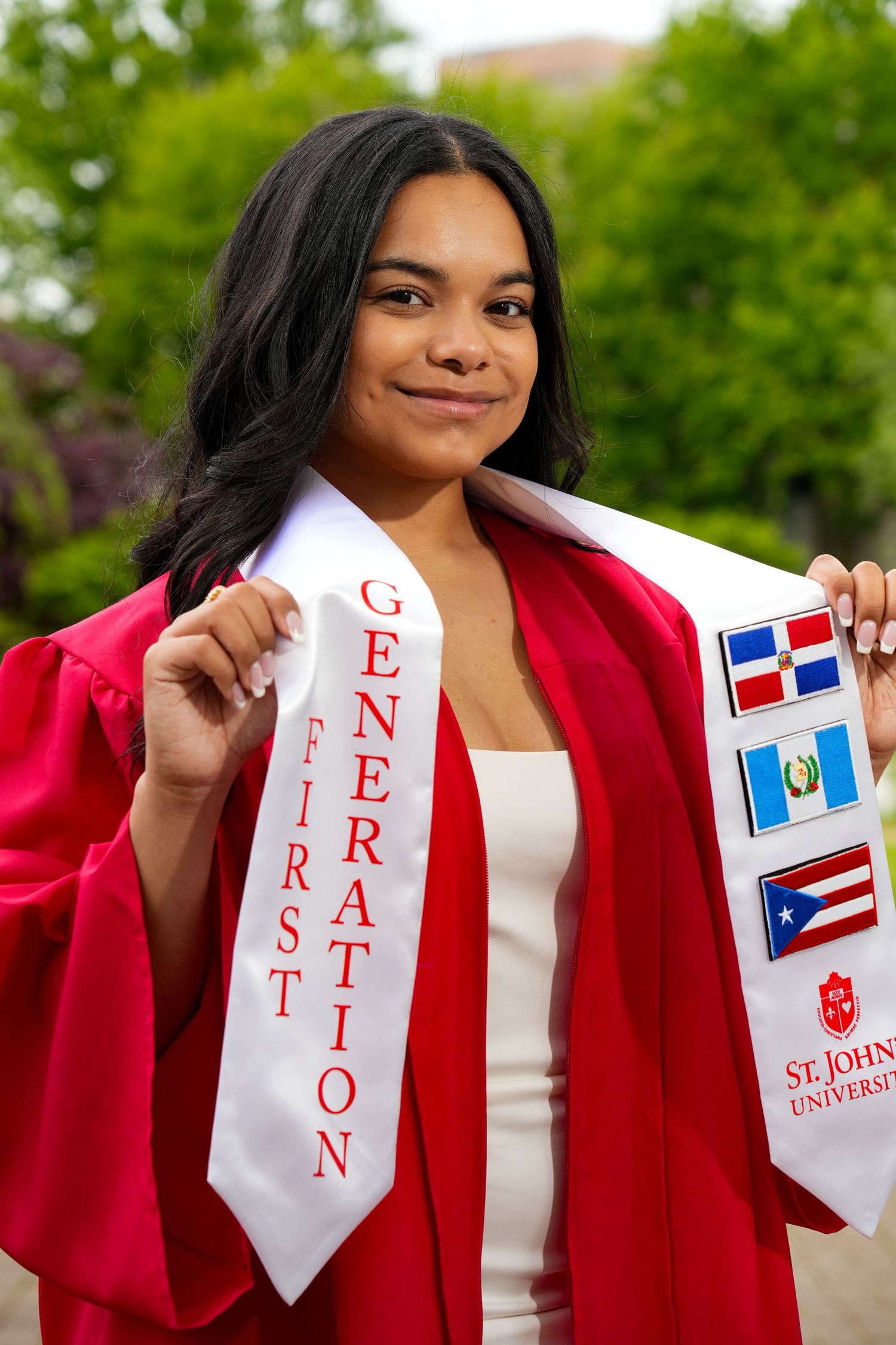 St. John's University first-generation graduate in red cap and gown holding sash with Dominican, Guatemalan, and Puerto Rican flags.