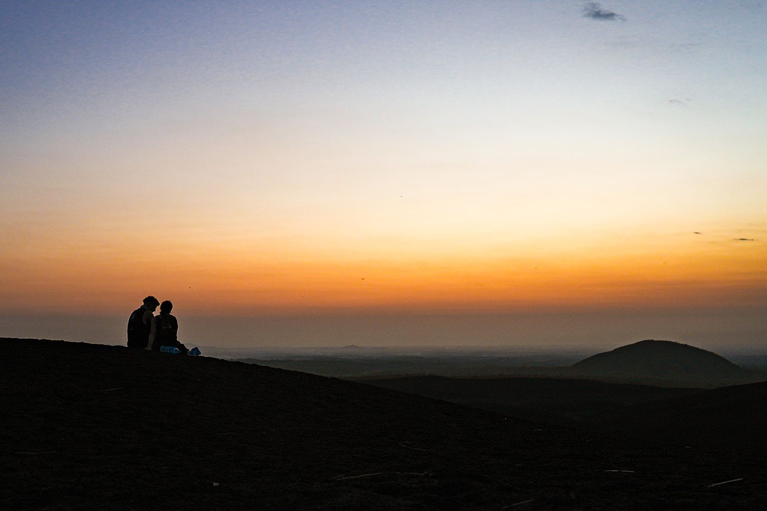 Silhouette of two people sitting together on a hilltop watching a vibrant orange and yellow sunset over distant mountains.