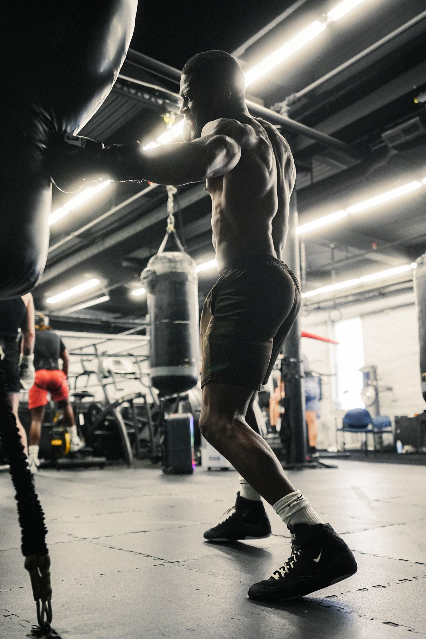 Muscular shirtless male boxer training on a heavy punching bag in a dark gym wearing black shorts and Nike shoes.
