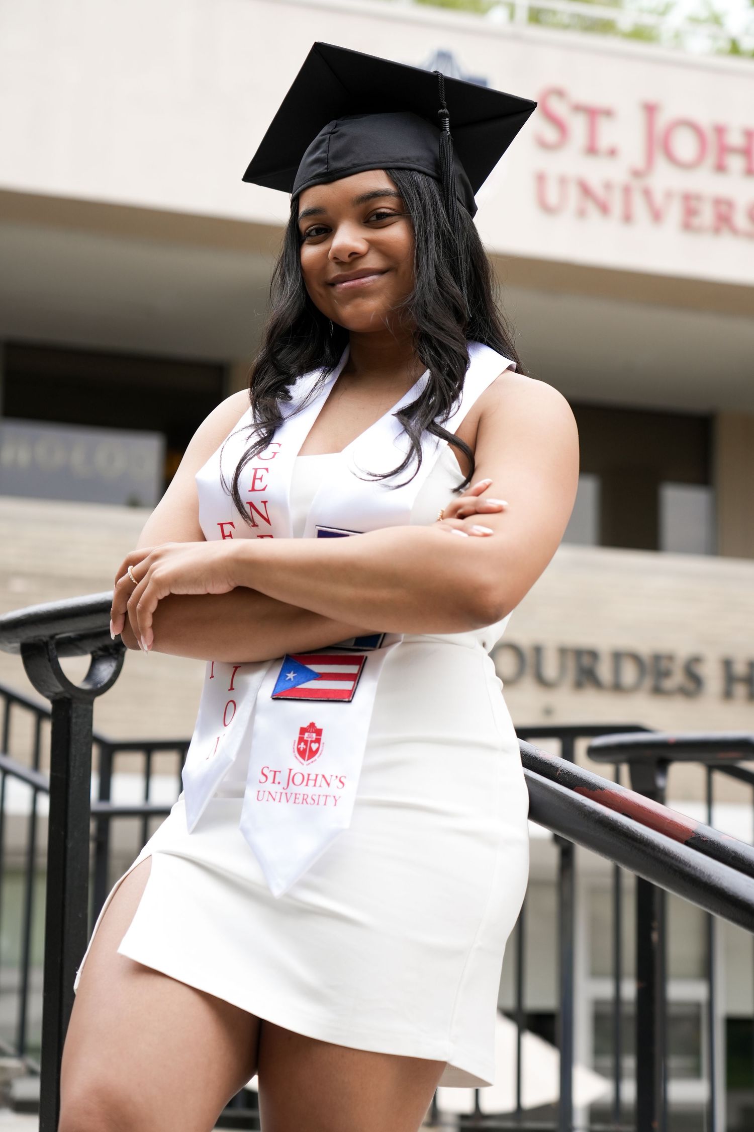 St. John's University graduate poses proudly in cap, gown sash, and white dress on campus steps.