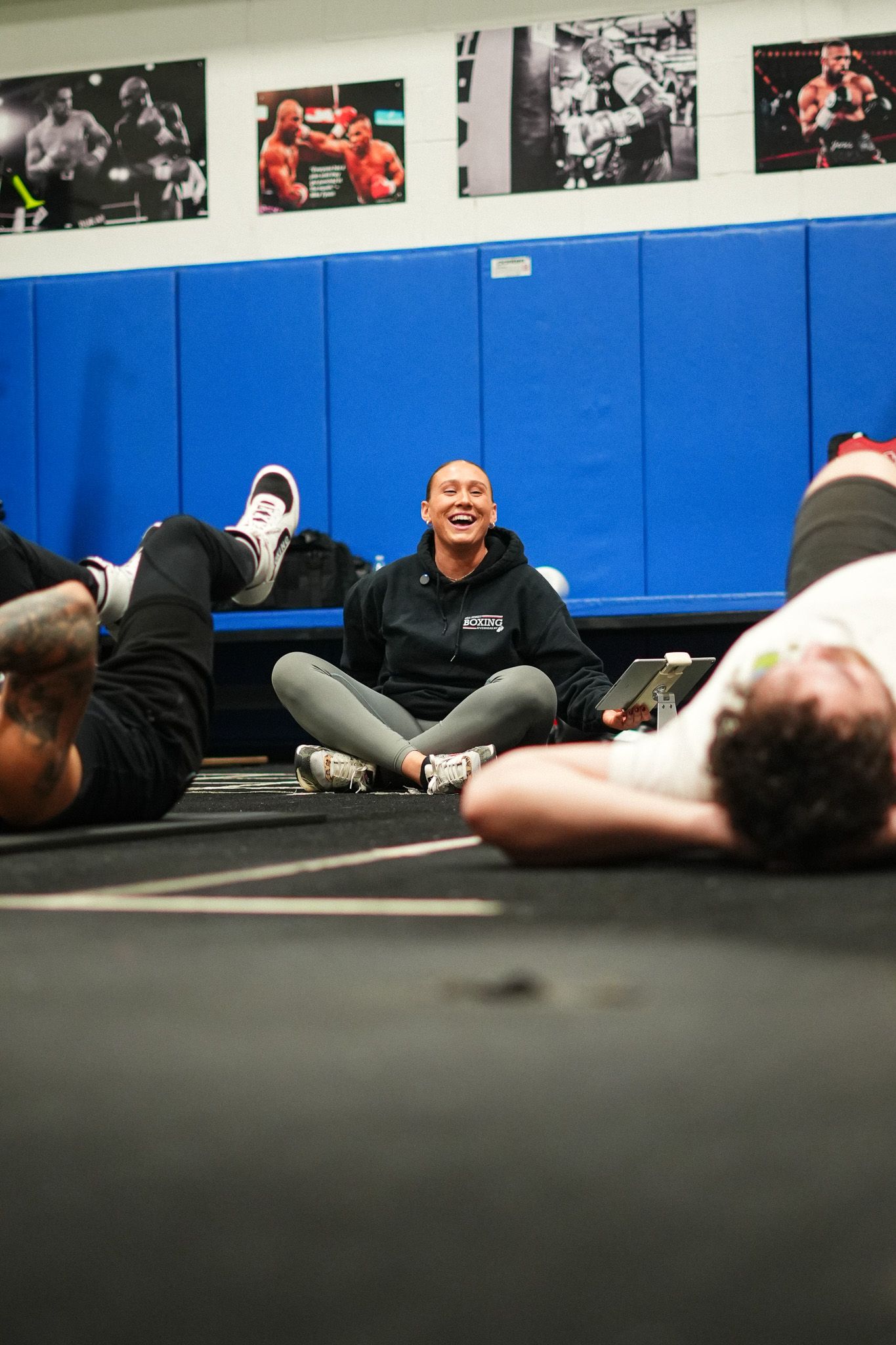 Athletes resting on gym floor during boxing training session with fight photos on wall.