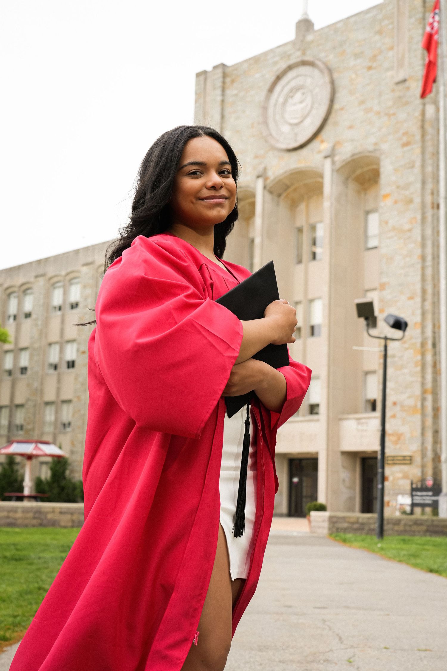 Young woman in red graduation gown holds cap outside university building, smiling proudly on graduation day.