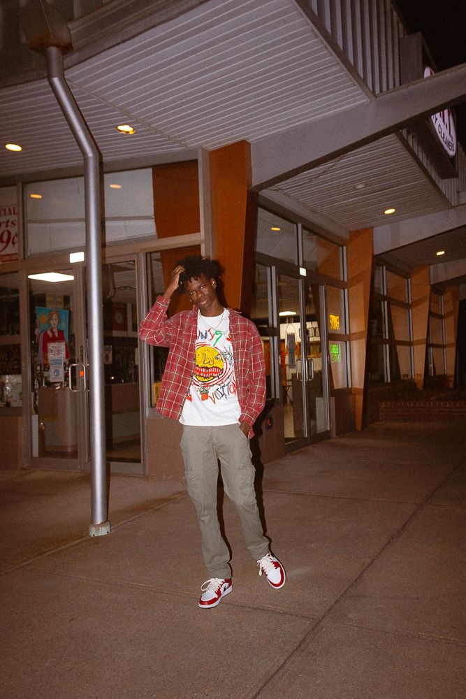 Young man in plaid shirt, graphic tee, gray pants, and red sneakers posing outside a store at night.