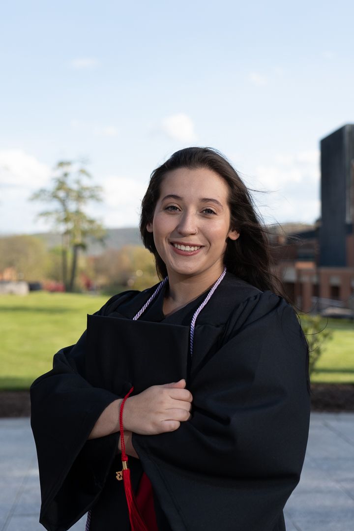 Smiling young woman in black graduation gown holding diploma outdoors on a sunny campus day.