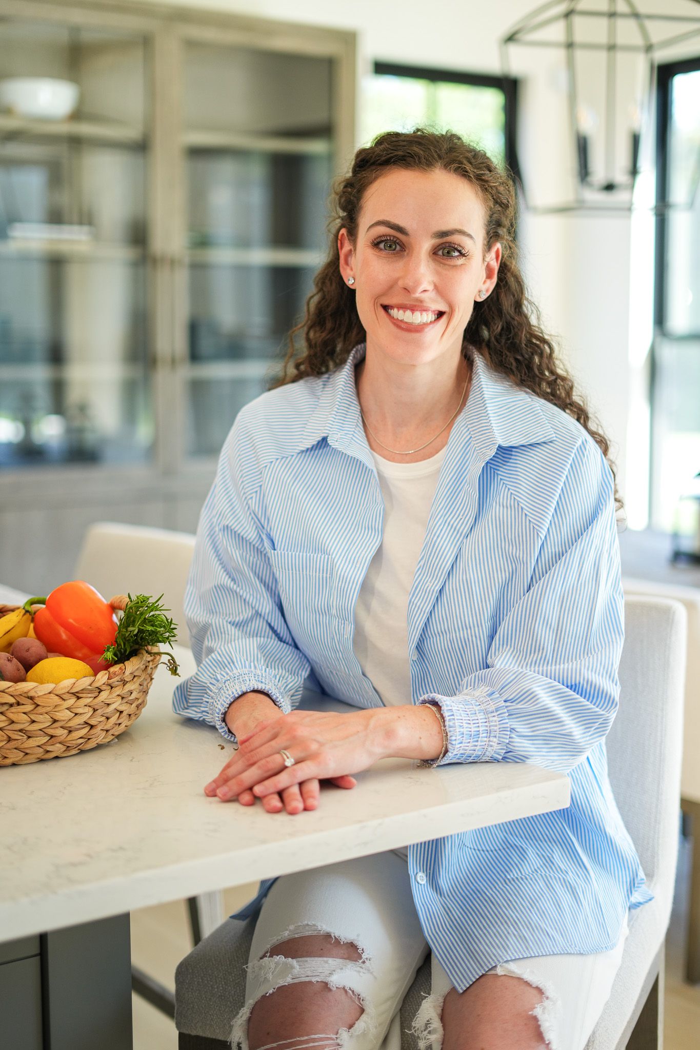 Smiling woman with curly hair in blue striped shirt sits at kitchen counter with fresh fruit basket.