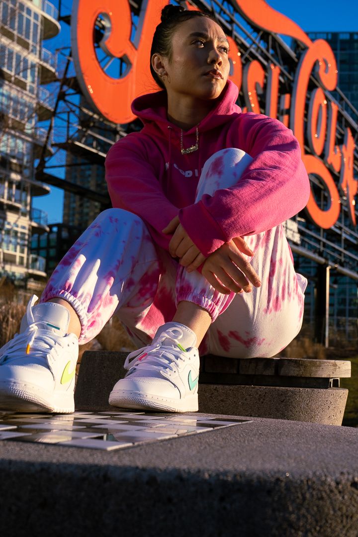 Young woman in pink hoodie and tie-dye pants sits on concrete steps wearing white sneakers near Pepsi-Cola sign.