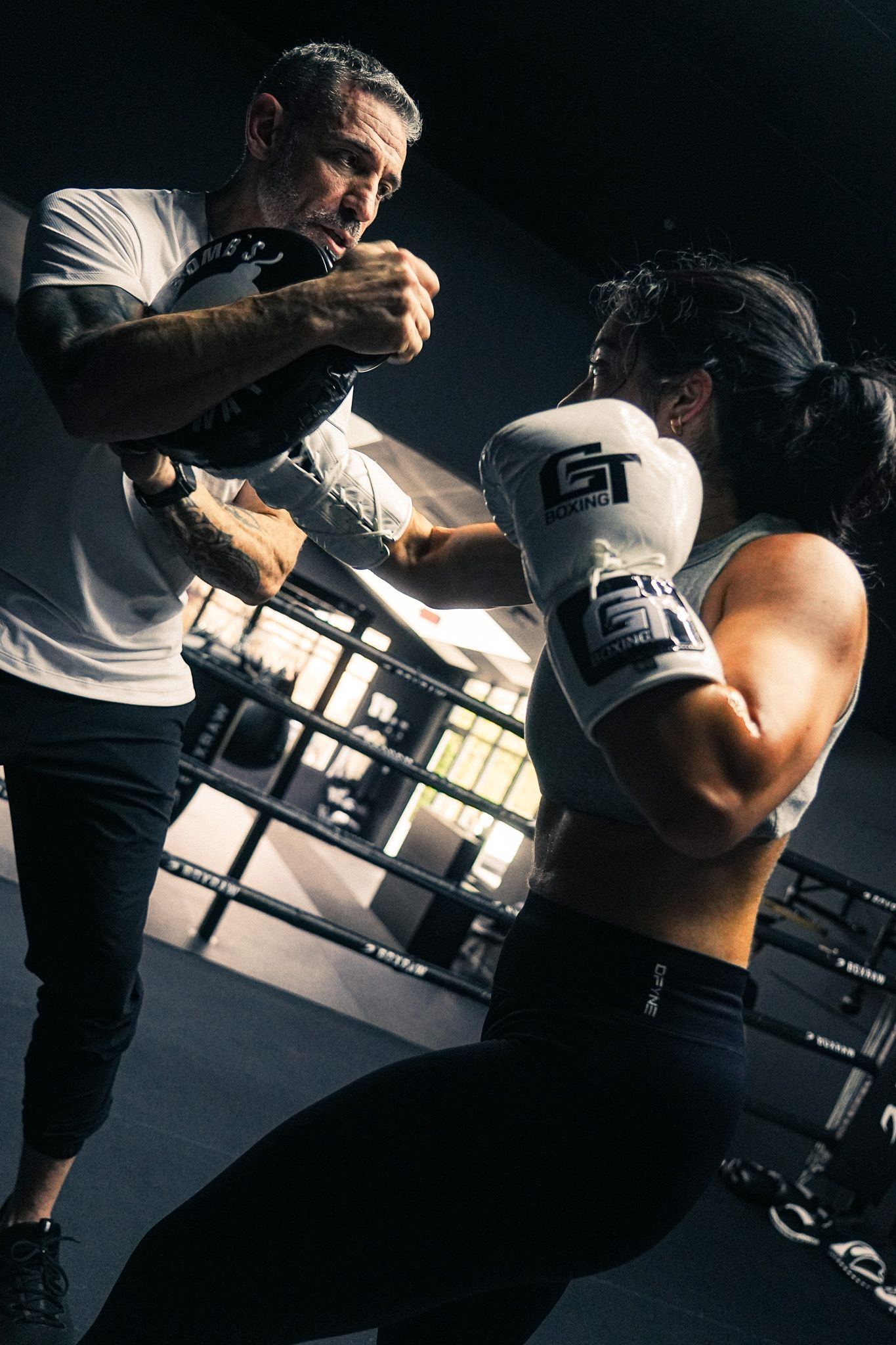 A woman in GT boxing gloves spars with a male trainer inside a dark professional boxing gym.