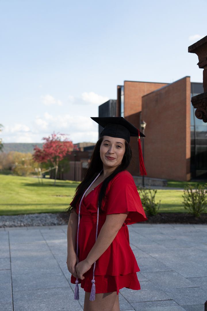 Young woman in red graduation cap and gown smiling on college campus with brick building background.