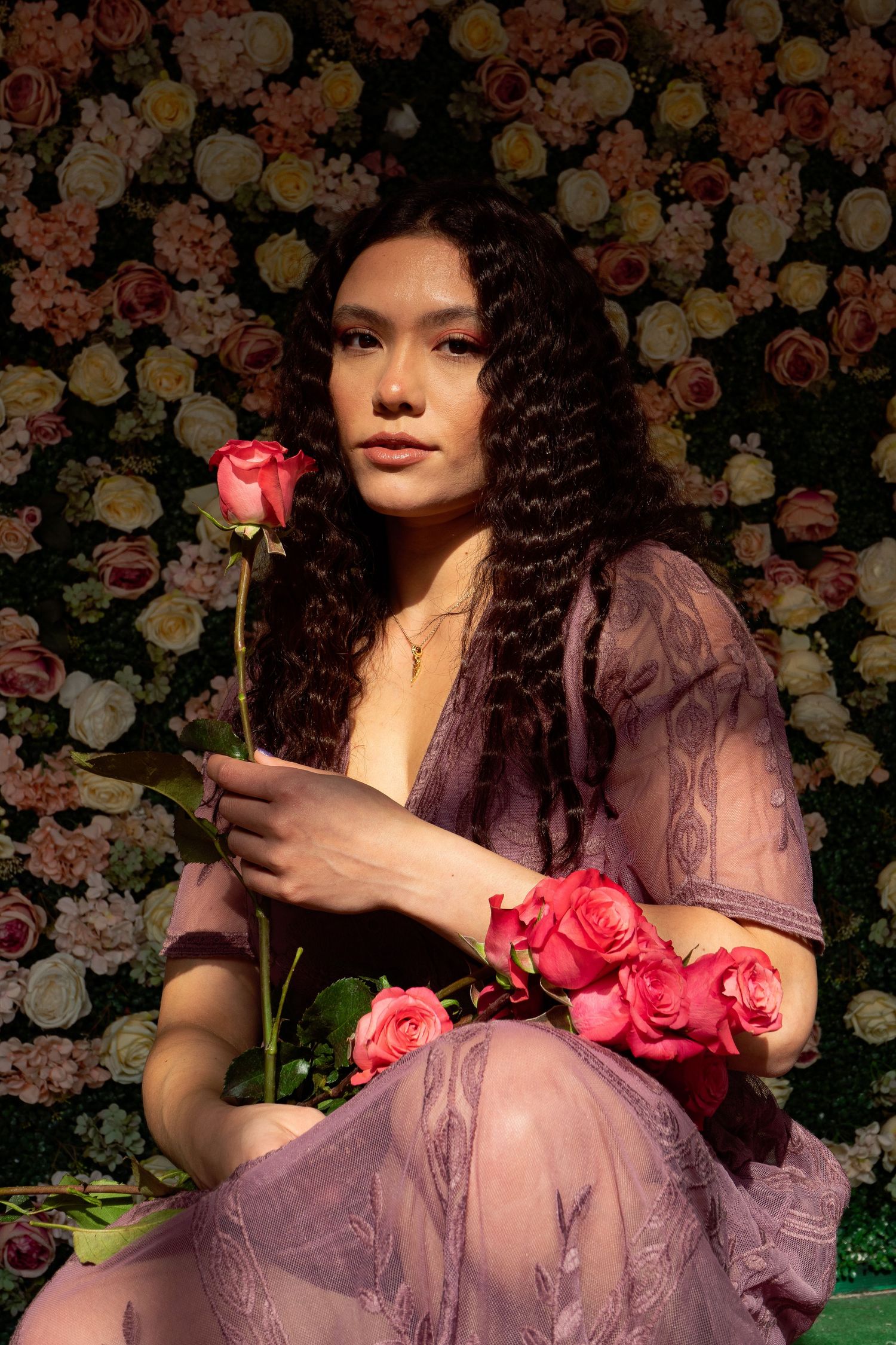 A woman in a purple lace dress holds pink roses against a floral wall backdrop.