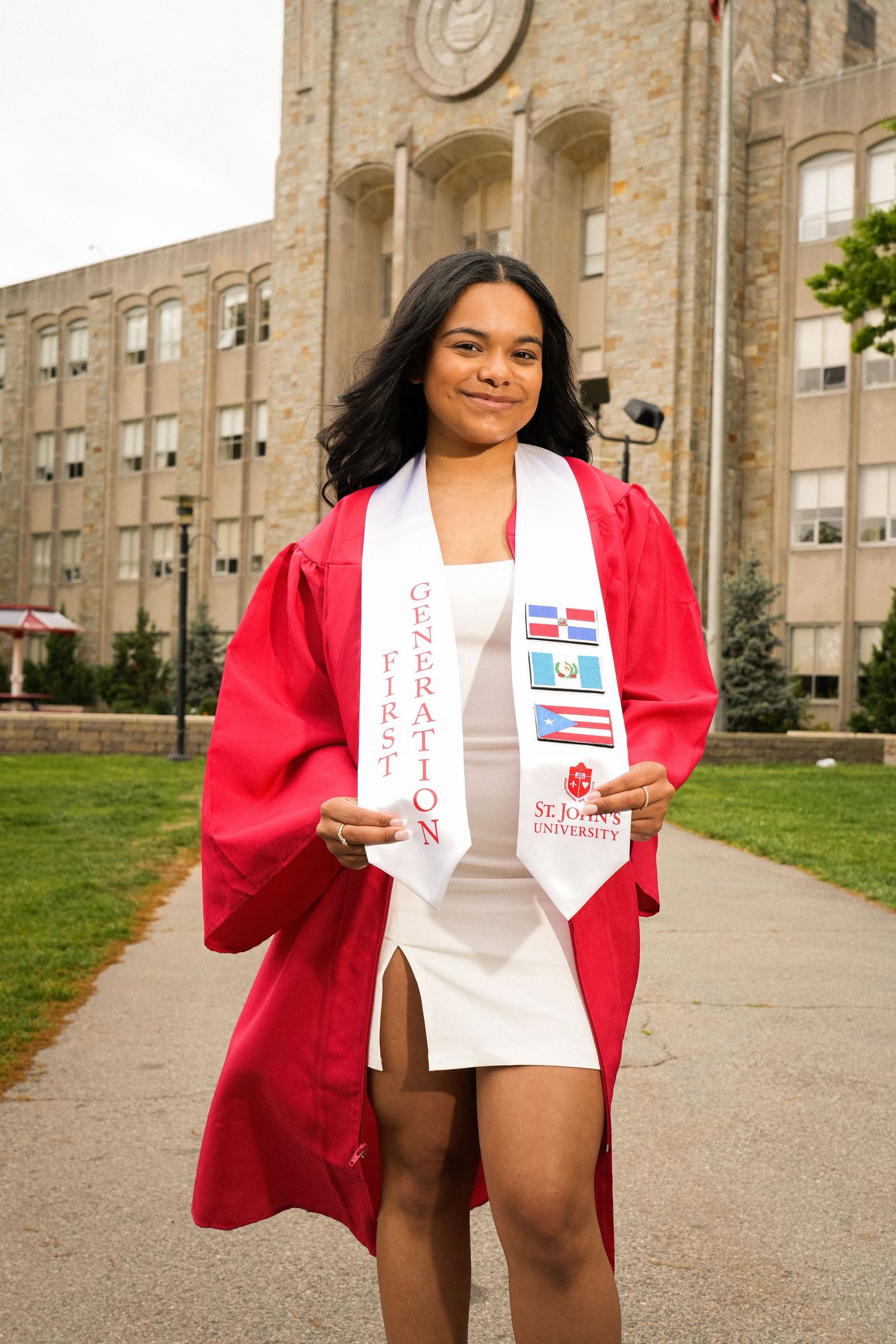 A smiling graduate in red cap and gown holds a First Generation stole with flags outside St. John's University.