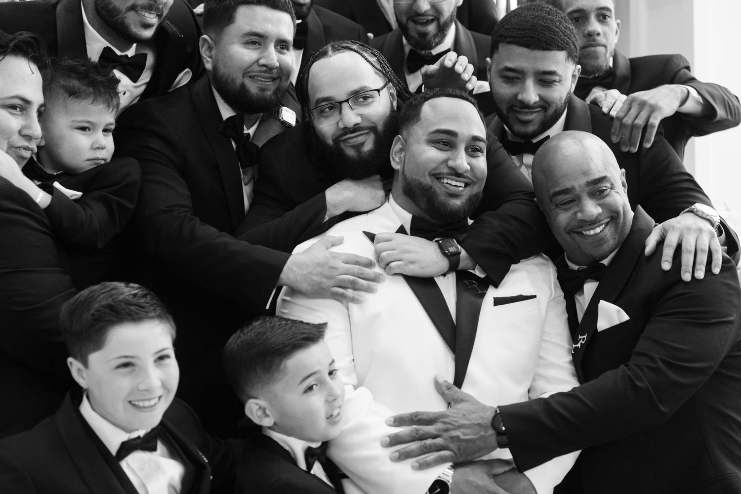 Black and white photo of a groom in white tuxedo surrounded by smiling groomsmen and children in bow ties.