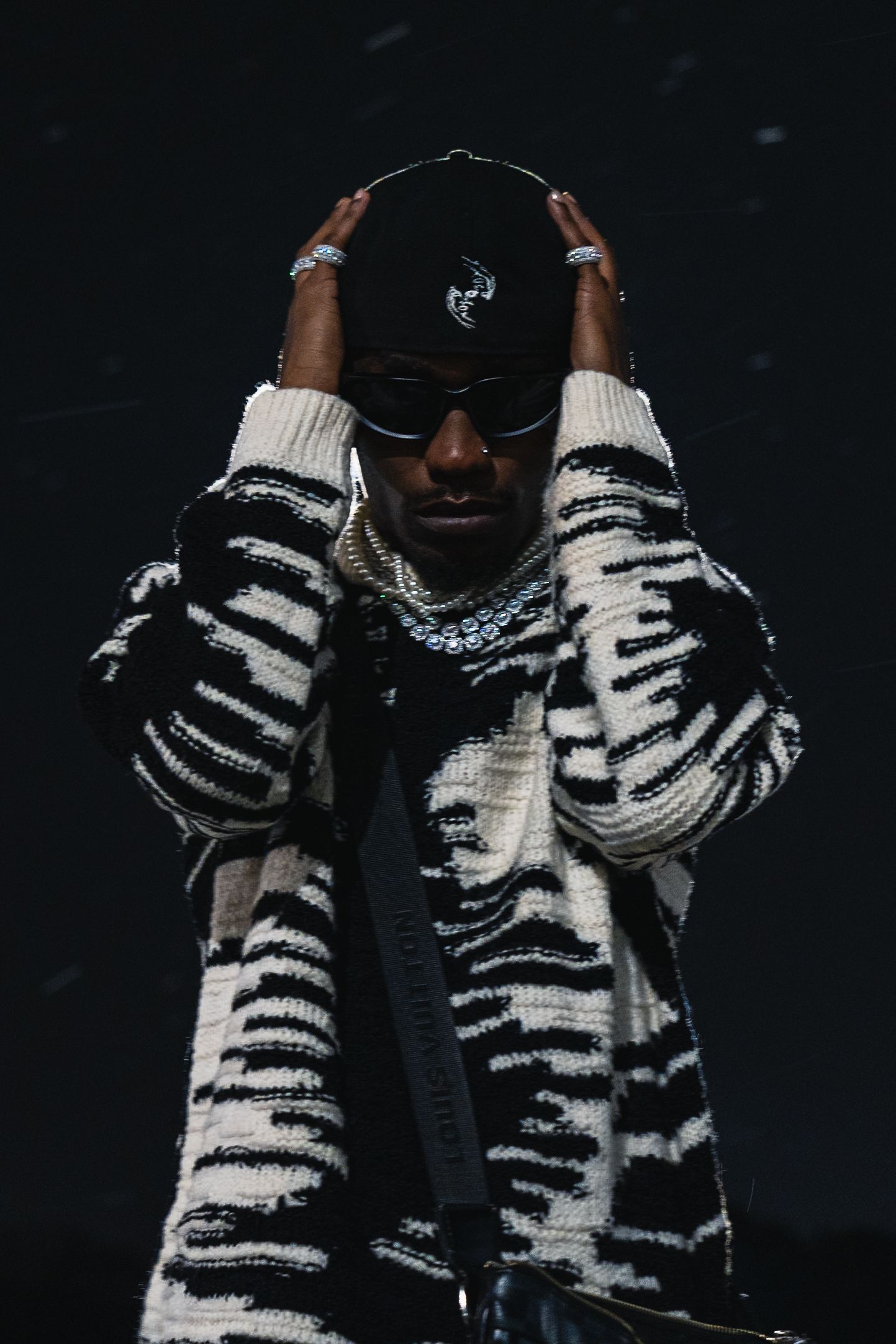 Person wearing black and white zebra print cardigan, sunglasses, and cap with diamond chain necklace posing dramatically.
