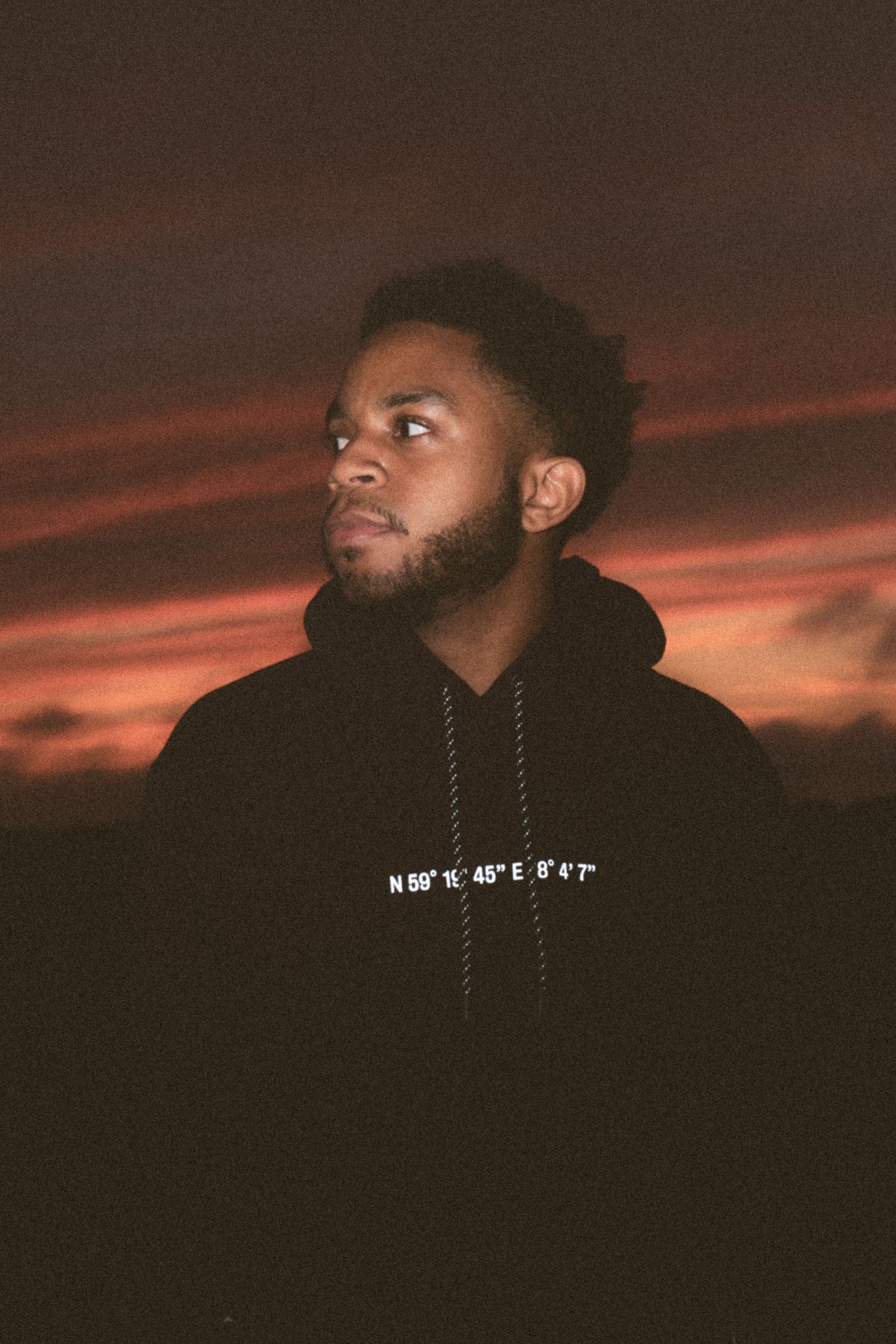Dramatic sunset portrait with person in dark hoodie against orange sky.