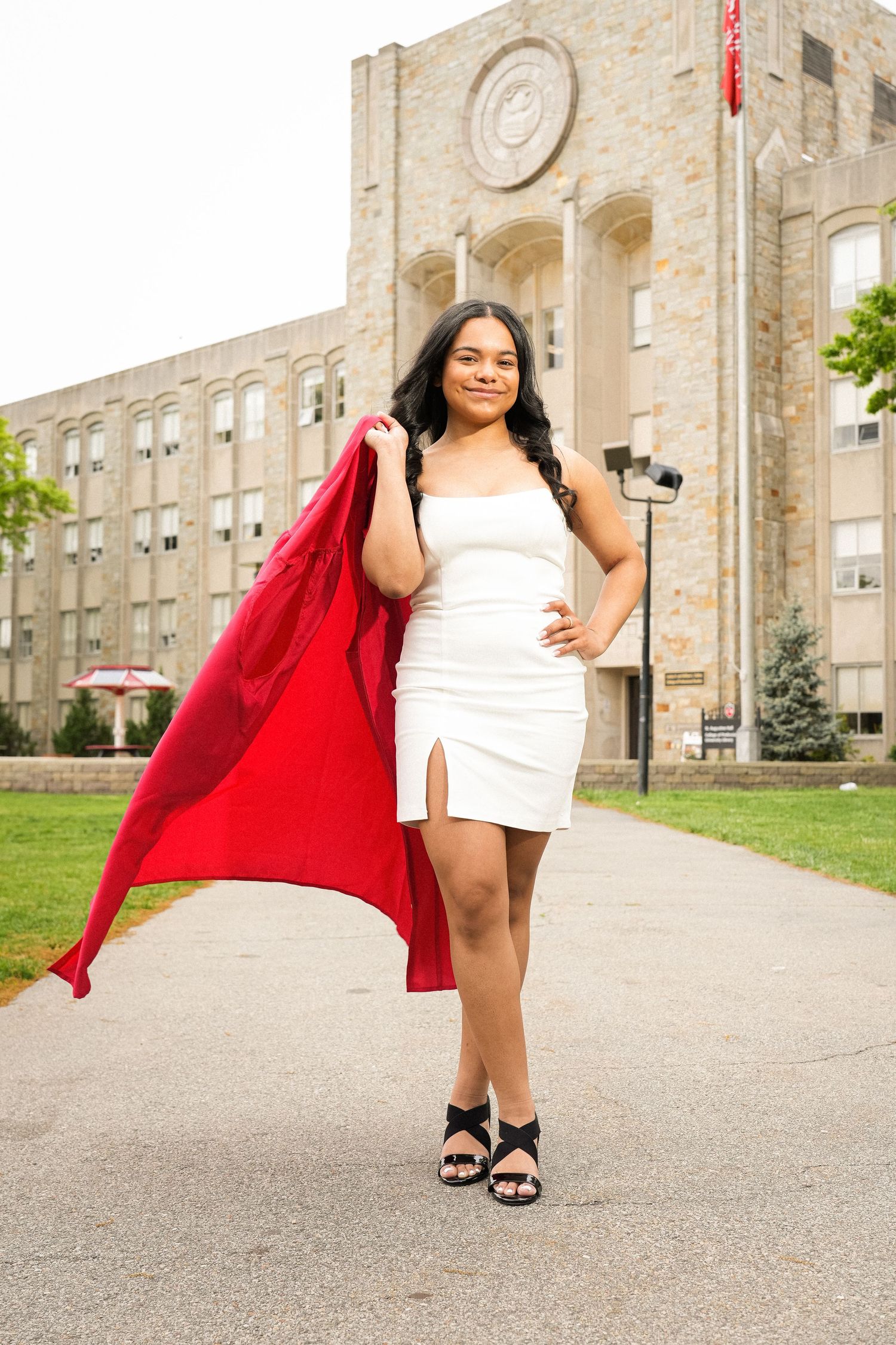 Young woman in white dress holding red coat poses confidently on campus pathway in front of a classic university building.