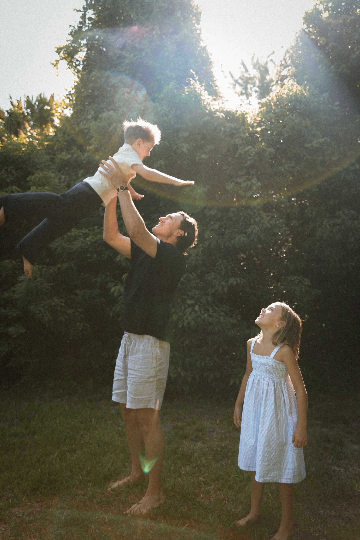 A father tosses a baby high in the air as a young girl laughs in a sunlit green backyard.
