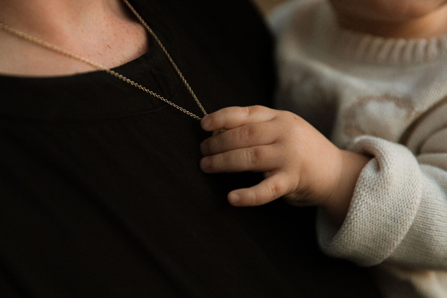 Close-up of baby hand grasping mothers gold chain necklace against black shirt.