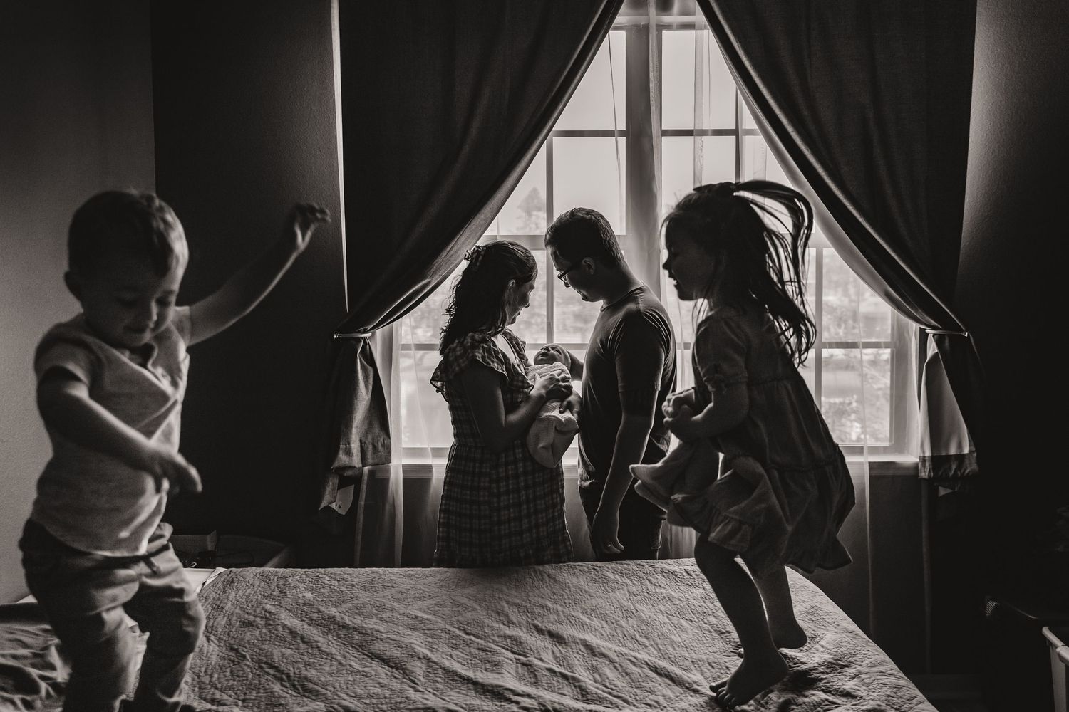 Black and white silhouette of children playing on a bed near a curtained window.