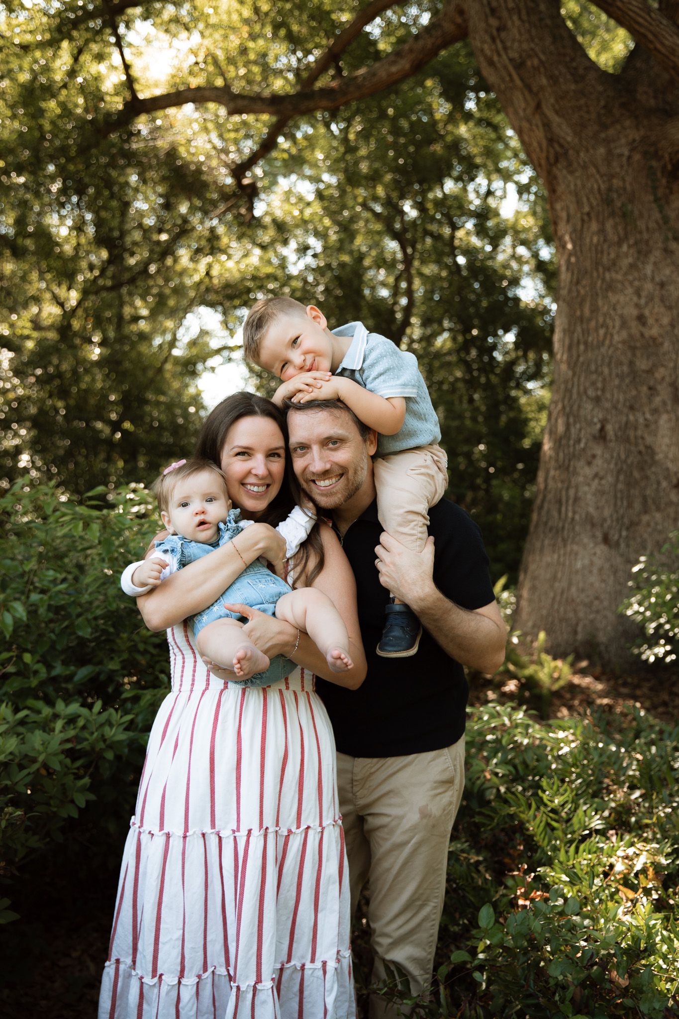 A smiling family of four poses in a sunlit green park, parents holding two young children.