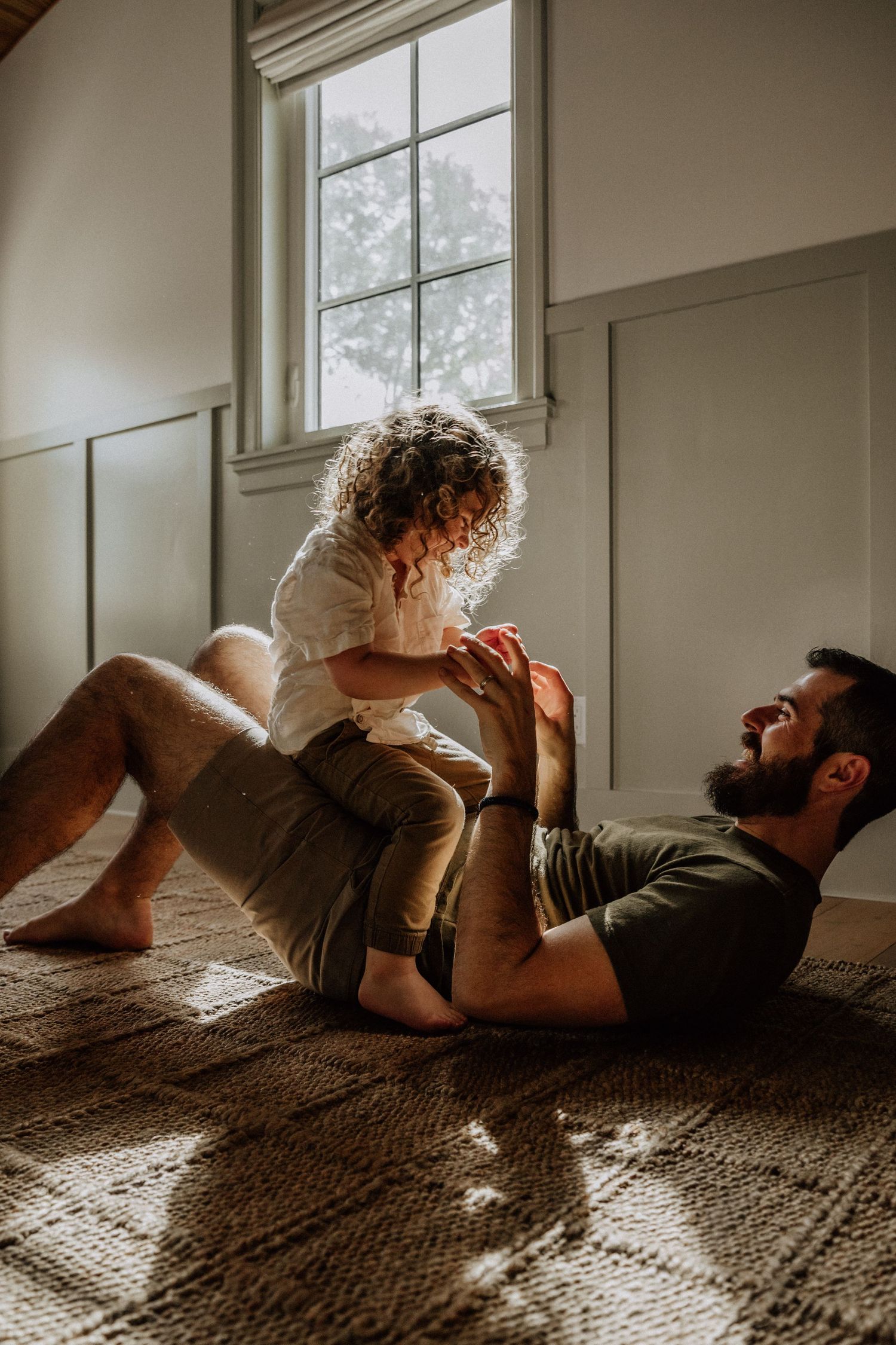 Laughing father playing with curly-haired toddler on floor in sunlit room.