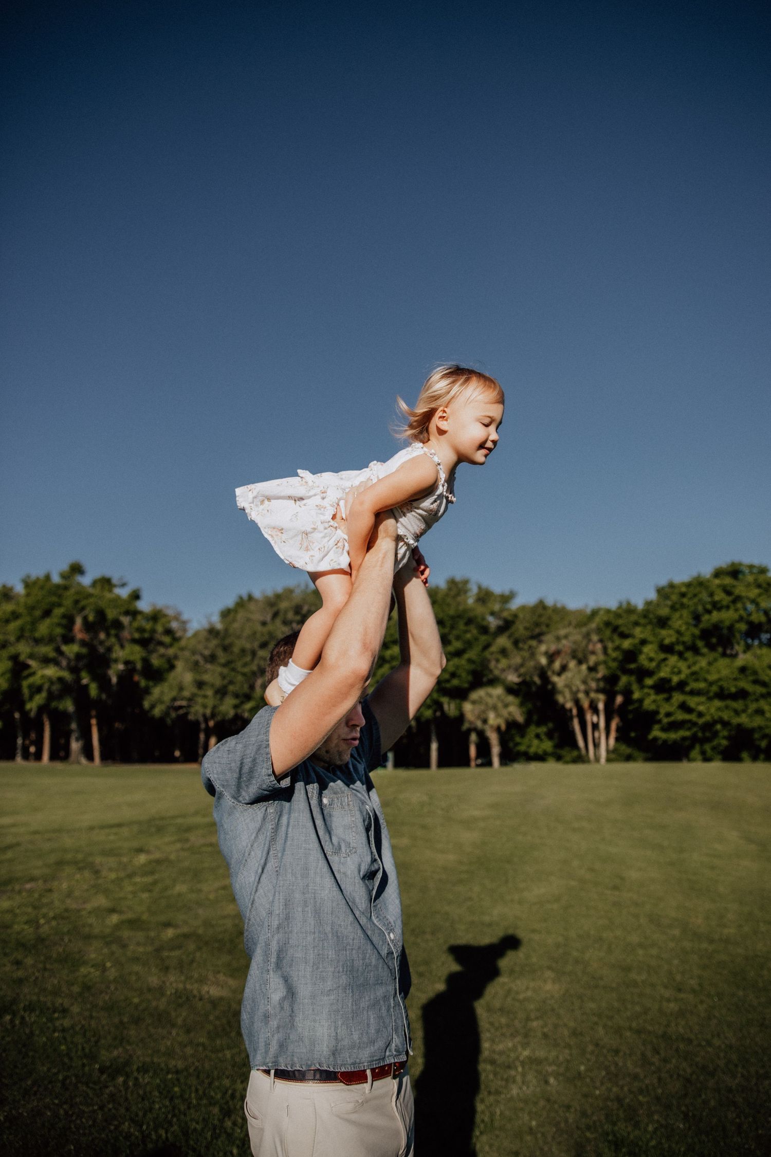 A father lifts his smiling toddler daughter high above his head in a sunny green park under a clear blue sky.