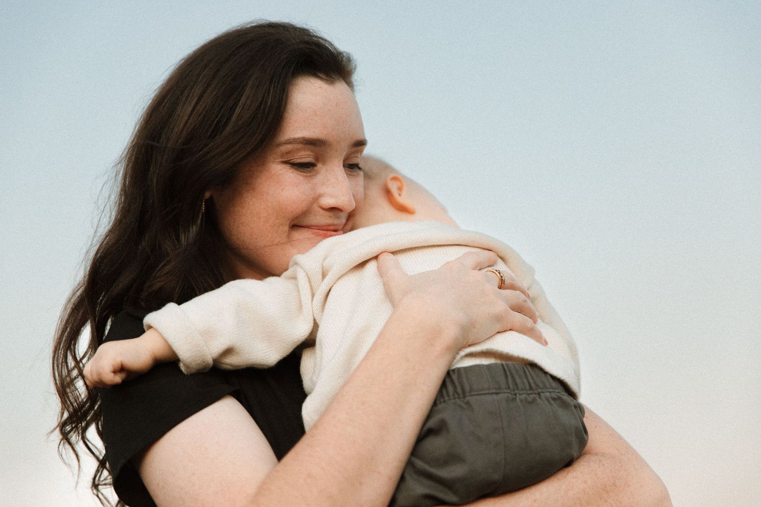 A smiling brunette mother lovingly embraces her infant child against a soft blue sky.