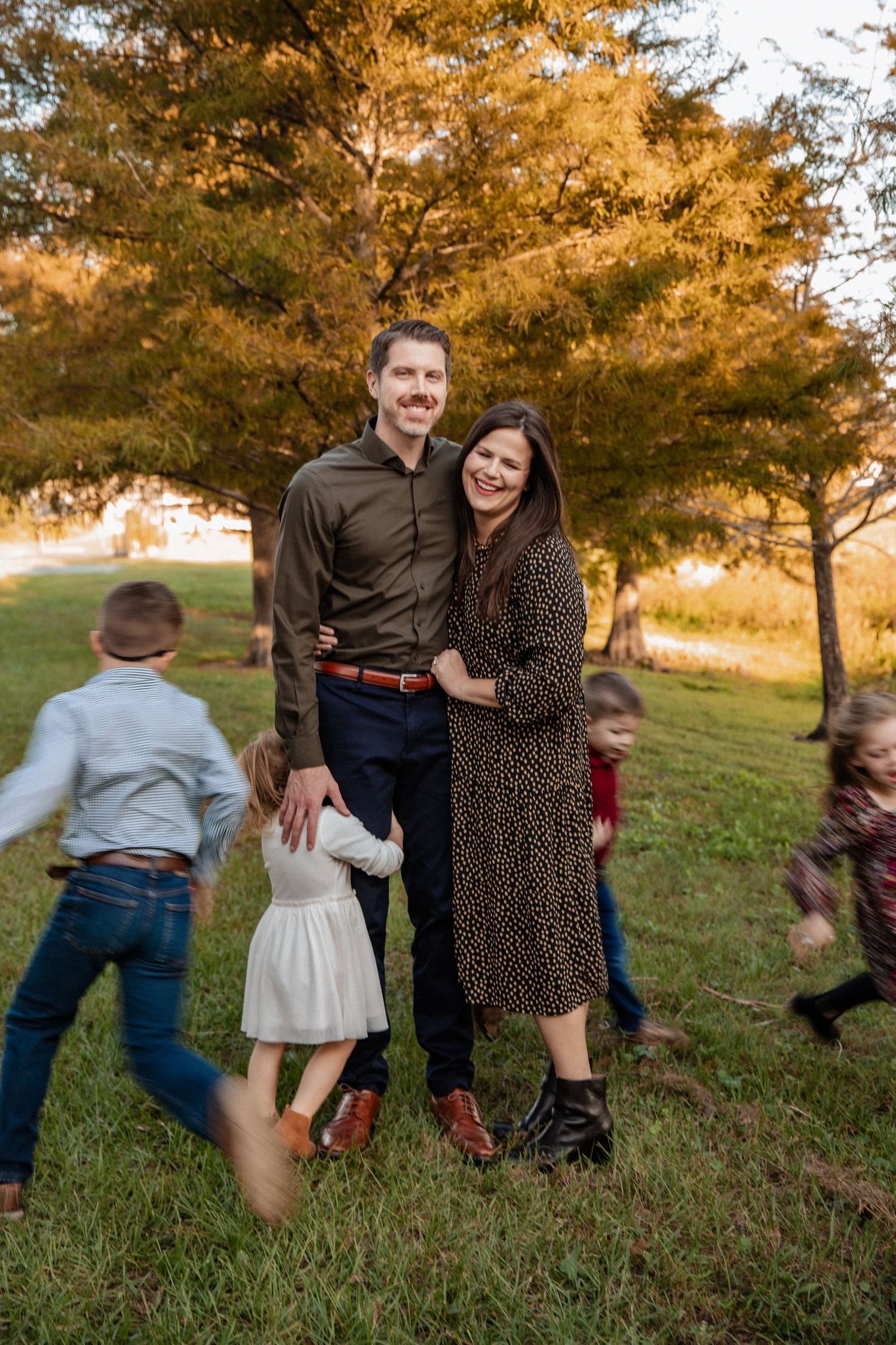 A smiling couple poses outdoors in autumn while their children play around them.