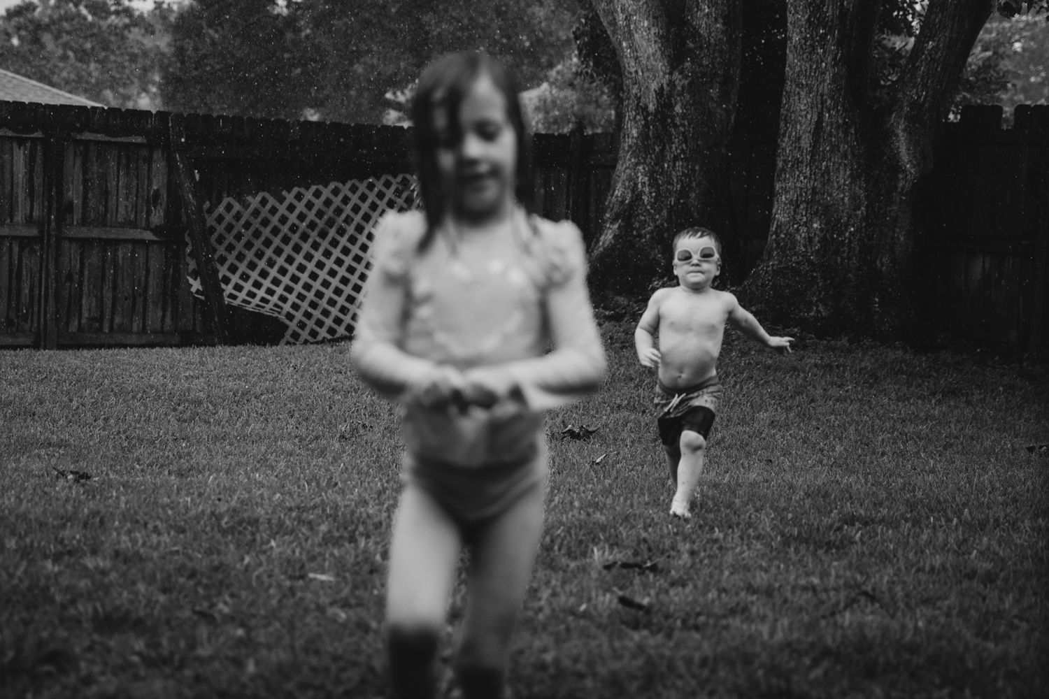 Black and white photo of two children playing and running in a backyard with a wooden fence.