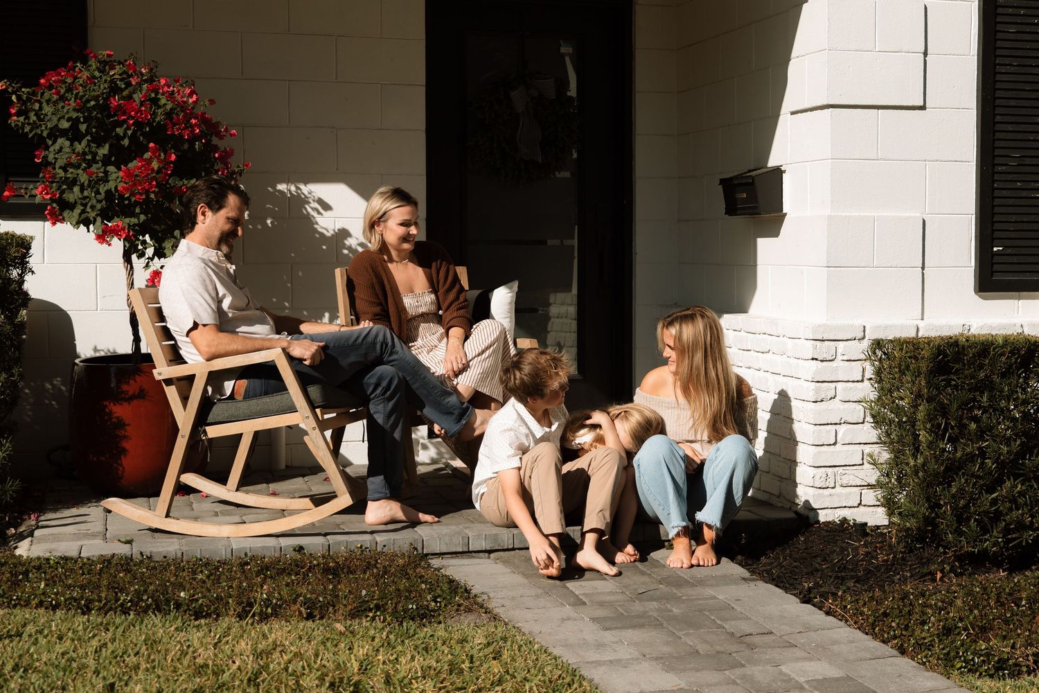 Happy family relaxing together on front porch of white brick home on sunny day.