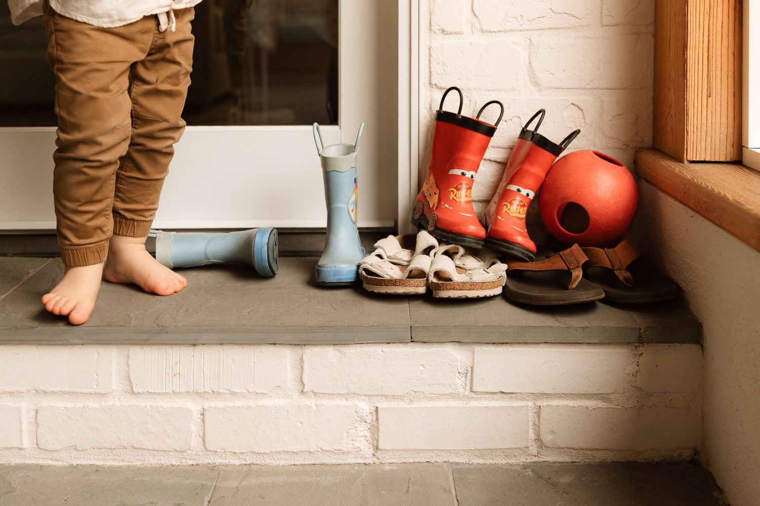 A barefoot child stands by a doorway next to a collection of colorful rain boots and sandals.