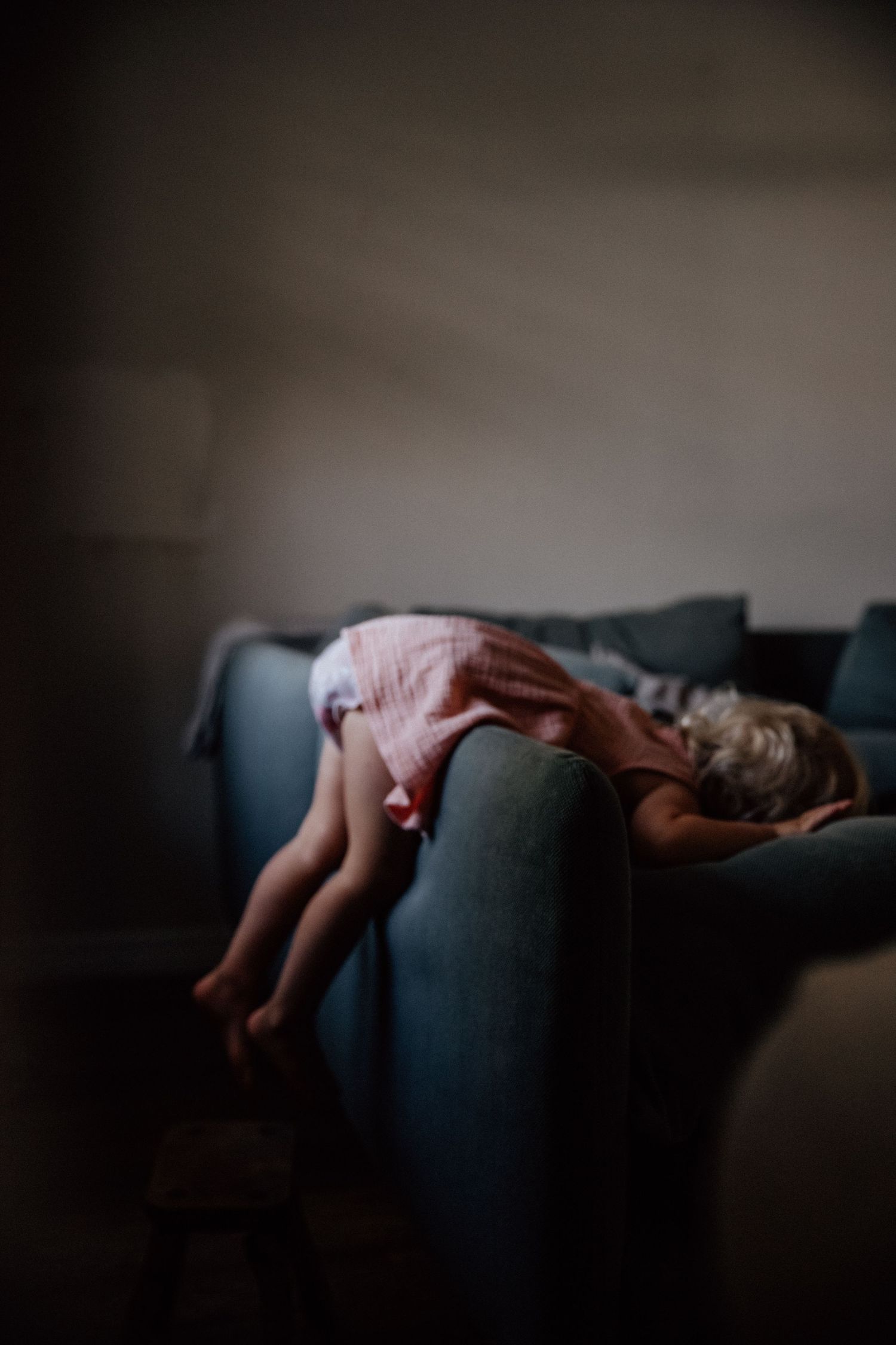 Young child in pink dress draped over back of blue sofa in dimly lit room.