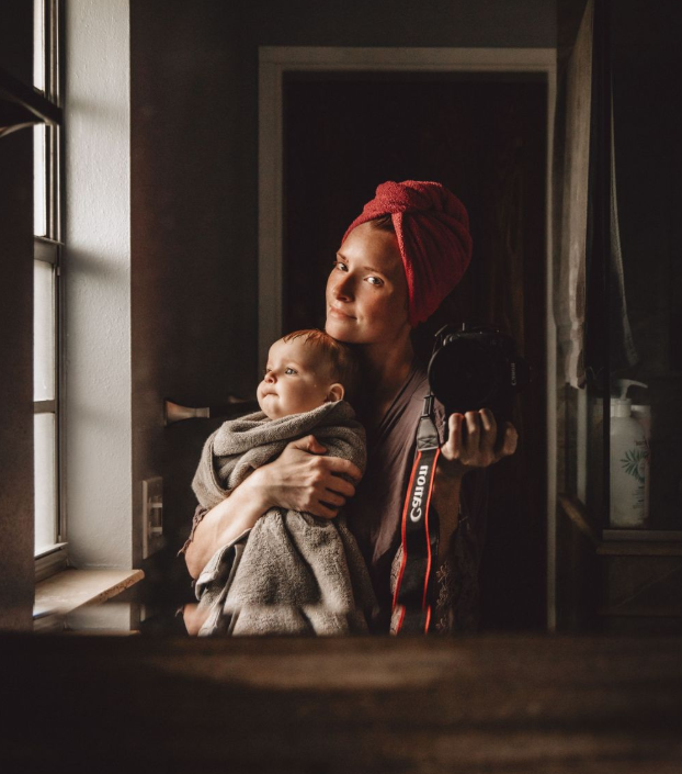 A mother wearing a red towel turban holds her baby while clutching a Canon camera near a window.