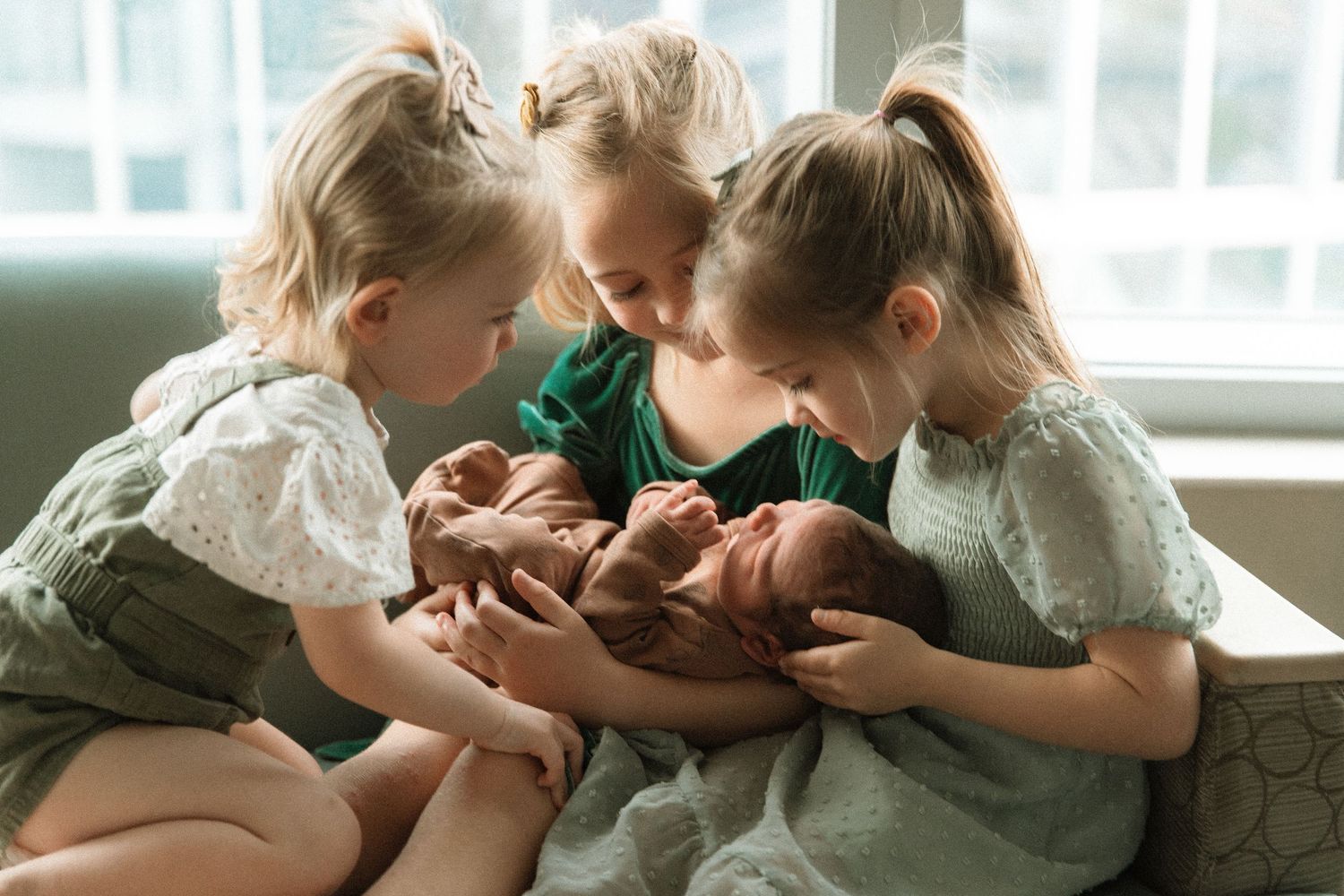 Three young sisters in sage green dresses tenderly holding newborn twins near a bright window.