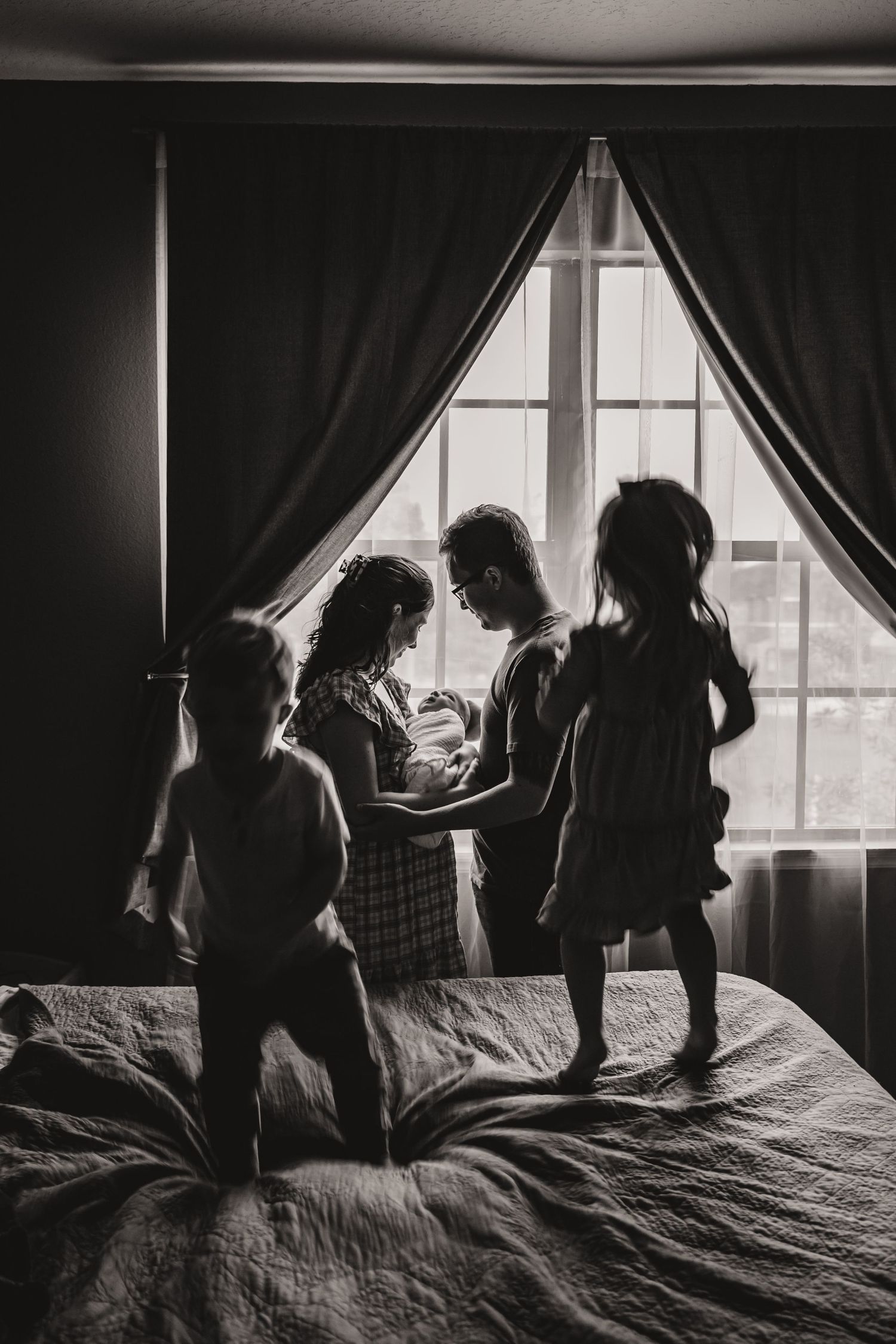 Black and white silhouette of family with children gathered around newborn by window.