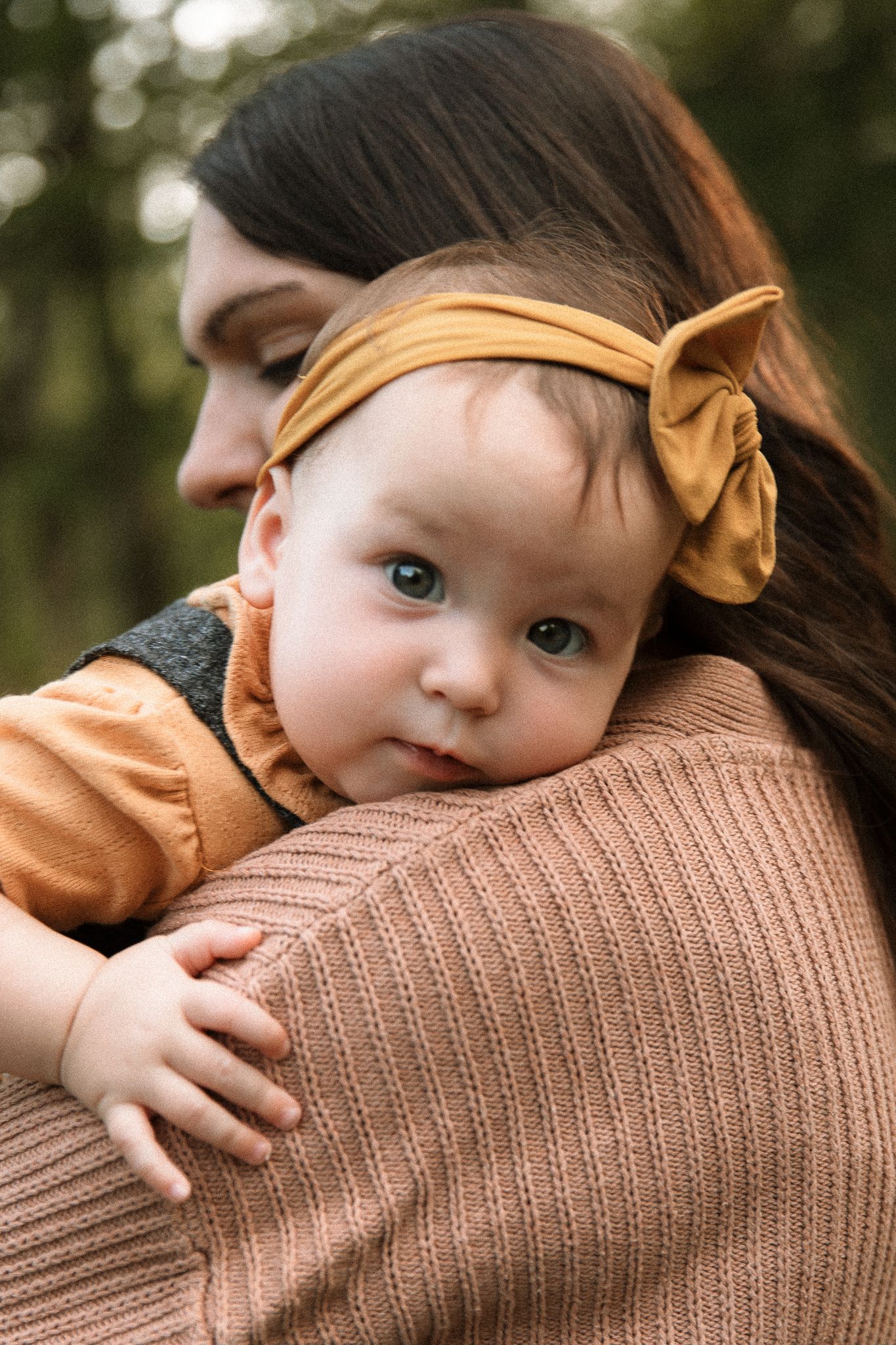 A mother with dark hair holds her blue-eyed baby girl wearing a mustard yellow bow headband outdoors.