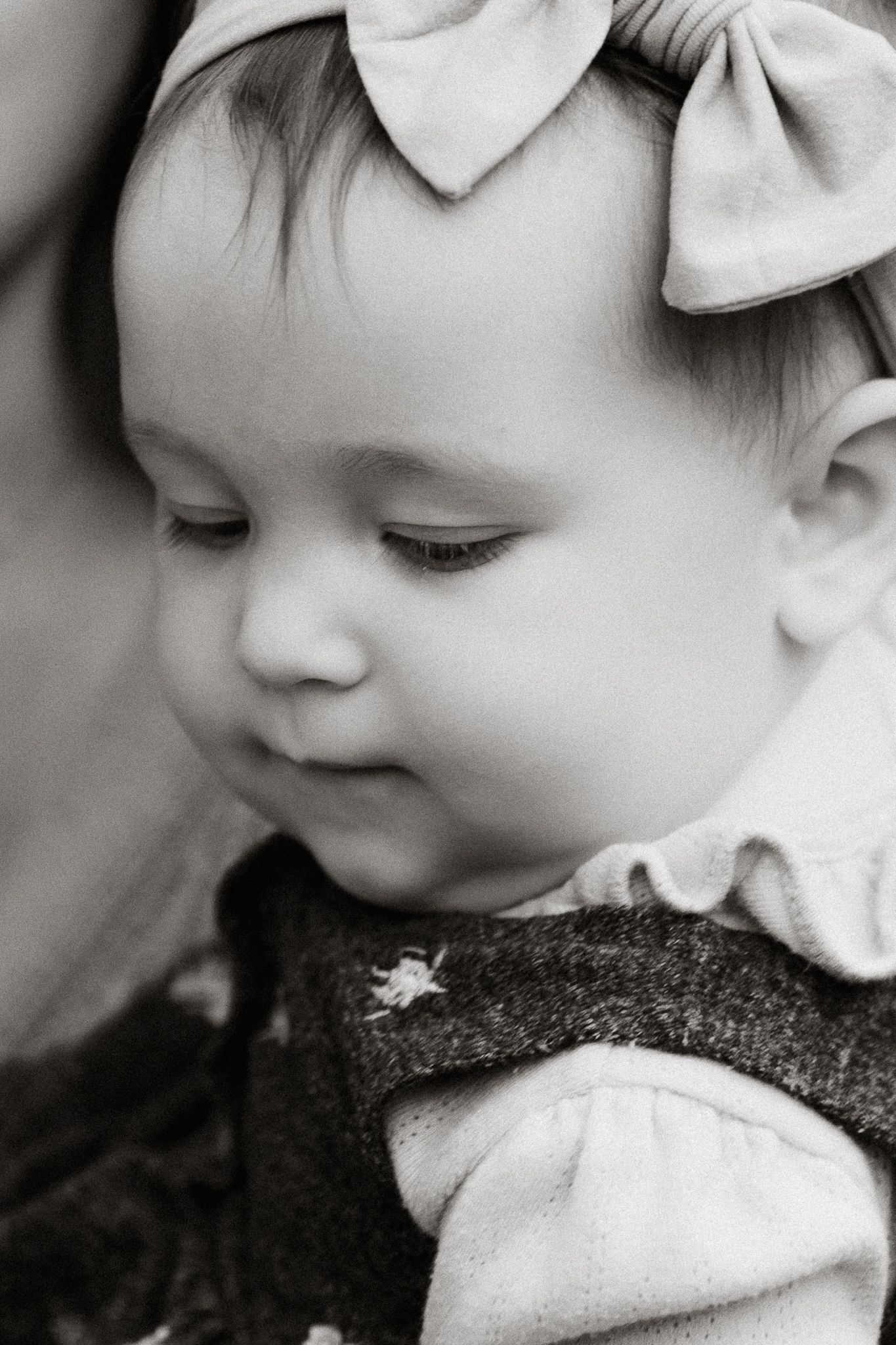 Black and white portrait of an adorable baby girl wearing a large bow headband looking down.