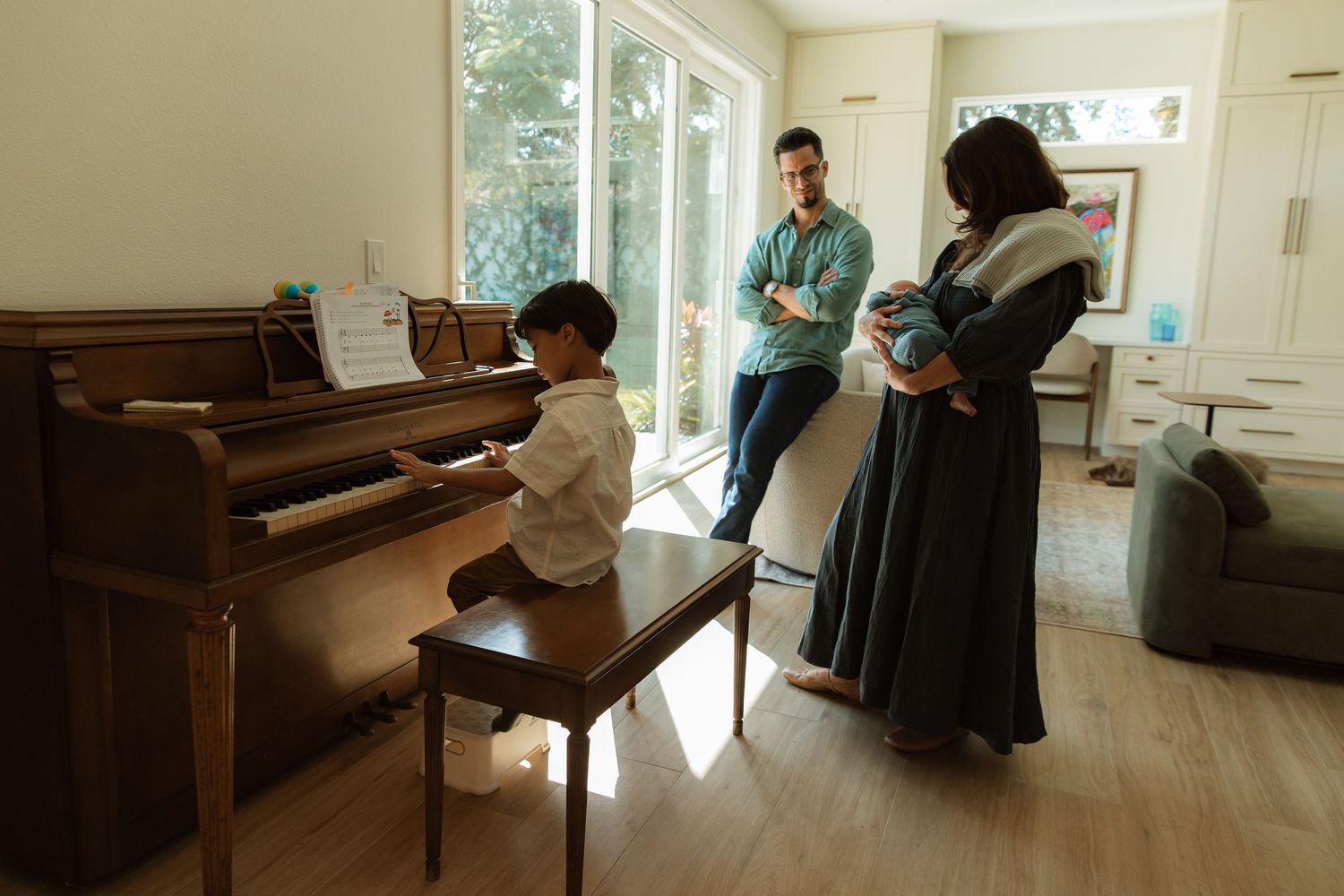 Family in living room as child plays upright piano while parents hold newborn baby.