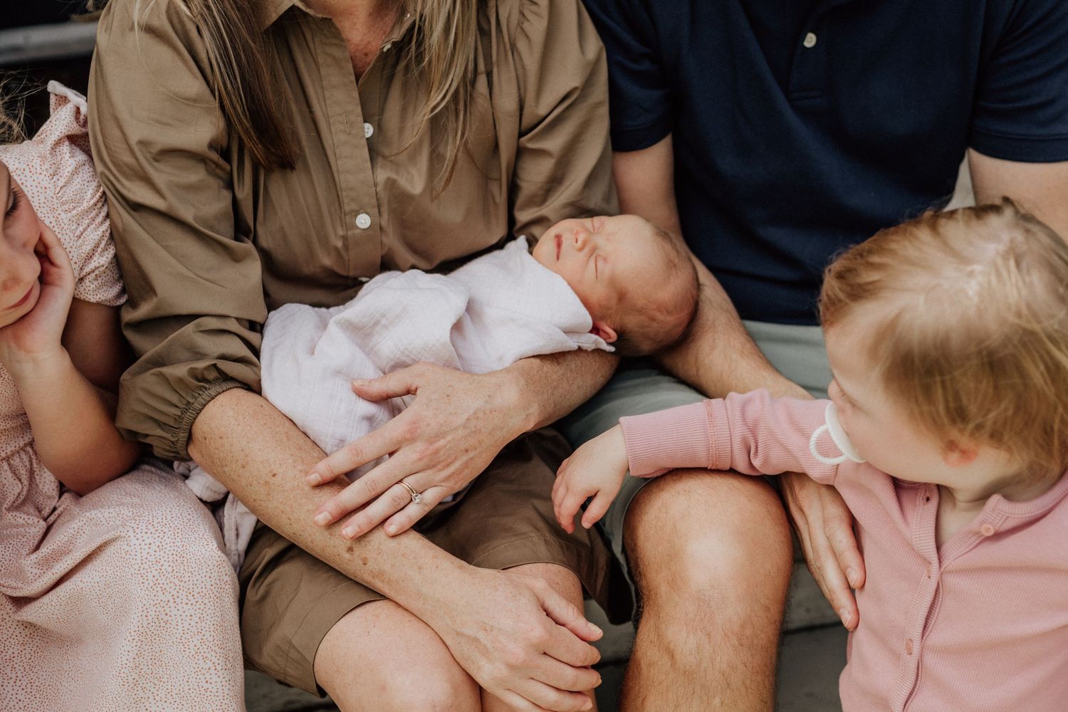 Family sitting together outdoors, parents cradling a newborn baby with two young children nearby.