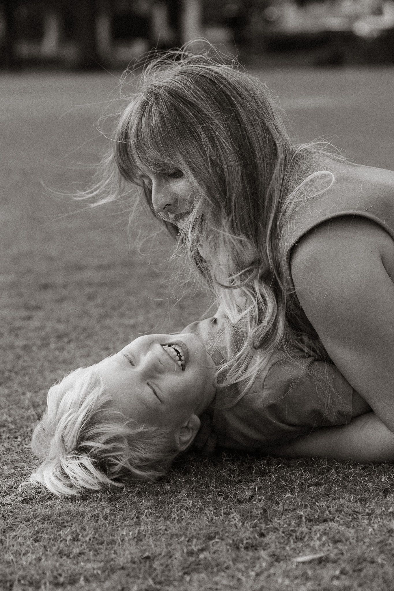 A black and white photo of a laughing child lying on grass while a woman leans over smiling.